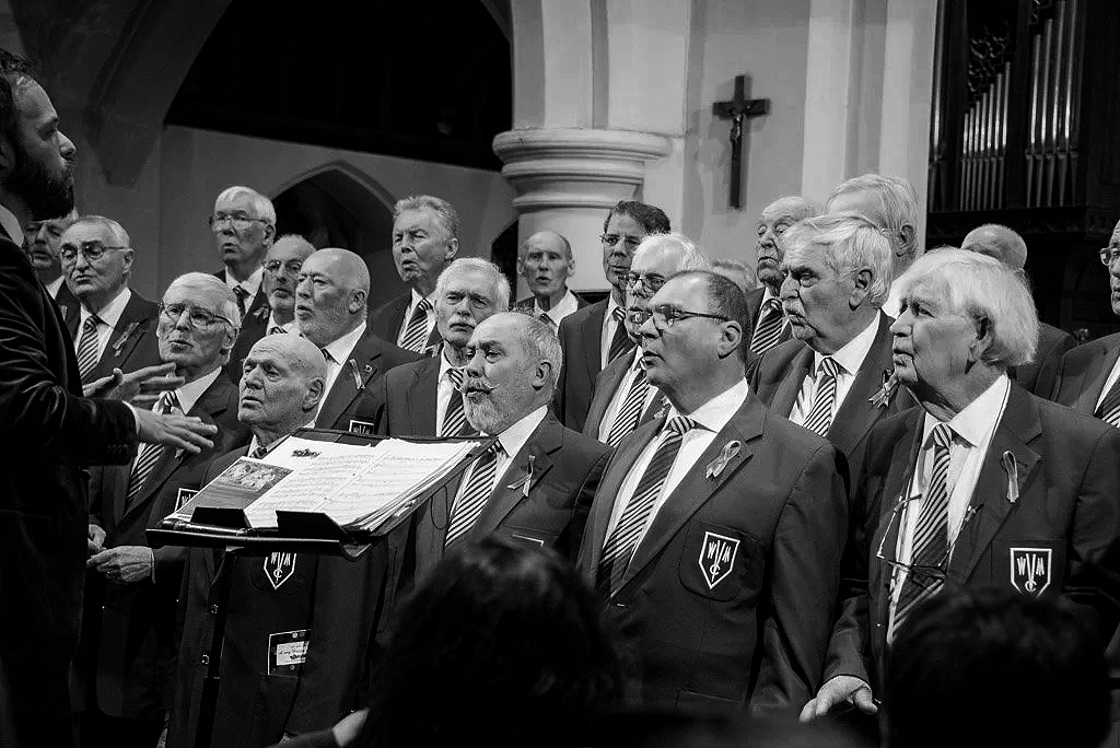 A choir of older men singing in a church, led by a conductor.