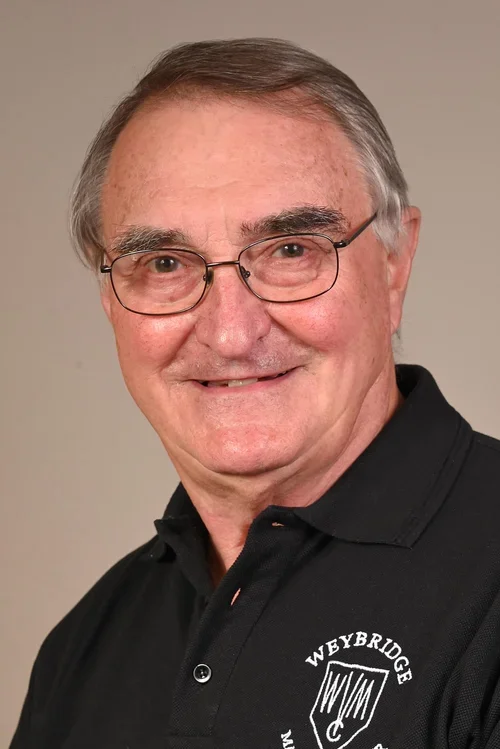 An older man with glasses, short gray hair, and a smile, wearing a black polo shirt with a 'Weybridge