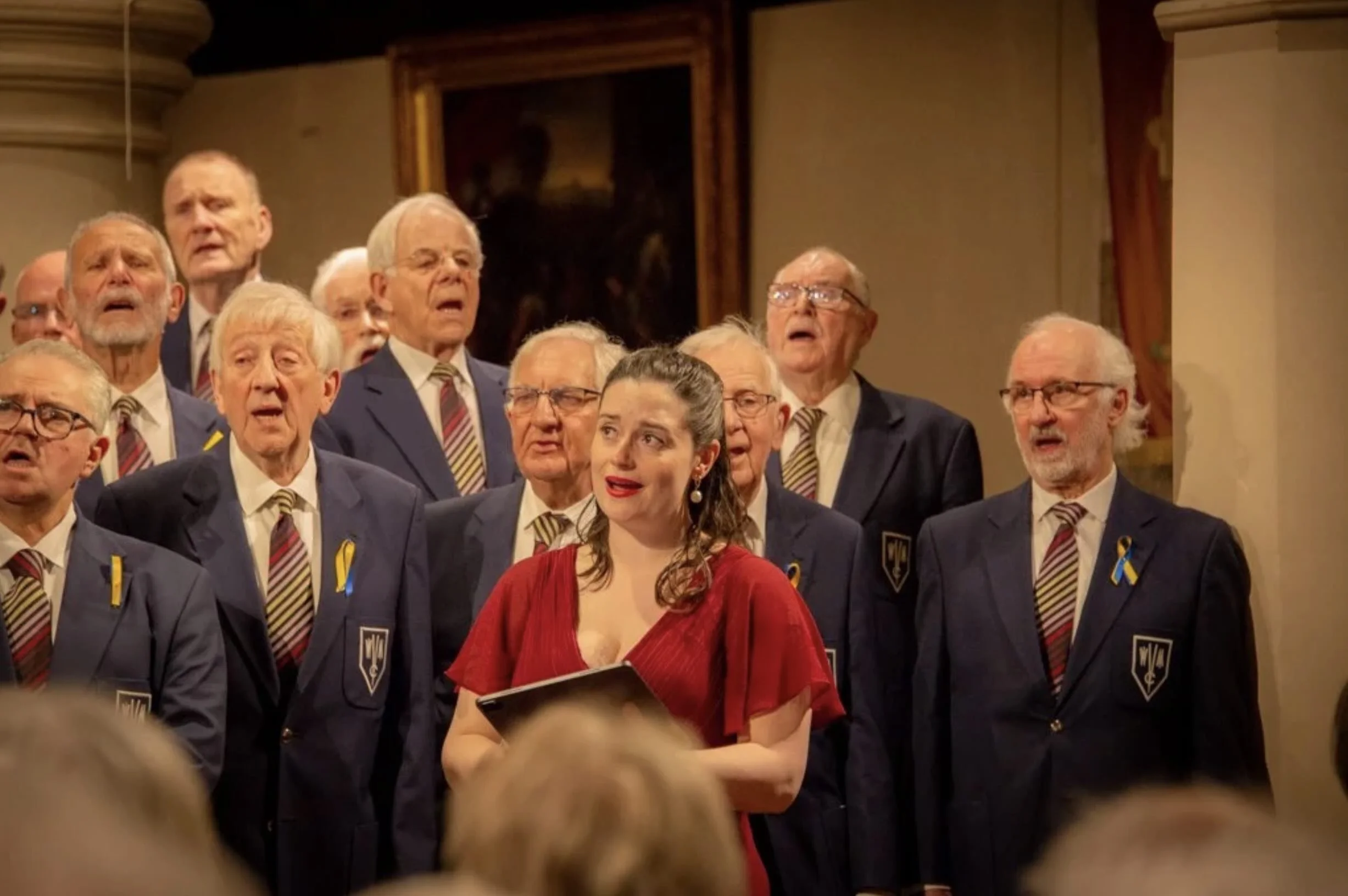 A woman in a red dress singing with a choir of elderly men in suits, wearing yellow ribbons, in a church or concert hall.
