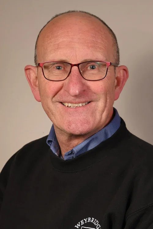 Portrait of a middle-aged man with glasses, wearing a black sweater over a blue collared shirt, smiling in front of a plain background.