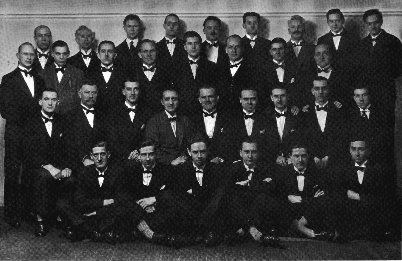A black and white photograph of a large group of men dressed in tuxedos, arranged in several rows, posing for a formal group photo indoors.