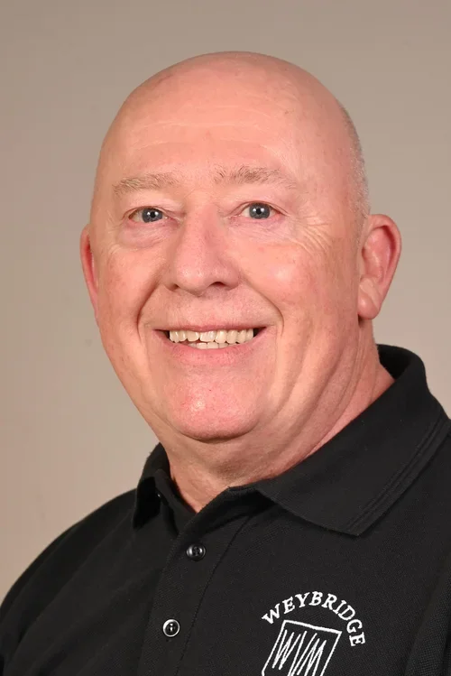 A smiling man with a bald head, blue eyes, and light skin, wearing a black polo shirt with a Weybridge logo.