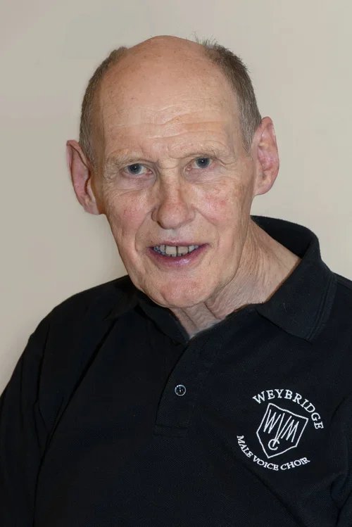 An elderly man with light skin, blue eyes, and short, thinning hair, wearing a black polo shirt with a "Weybridge Male Voice Choir" logo.