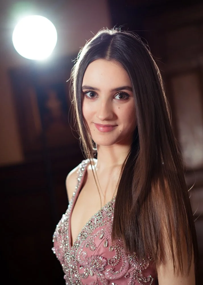 Young woman with long brown hair smiling, wearing a pink dress with intricate beadwork and embellishments, indoors with soft lighting