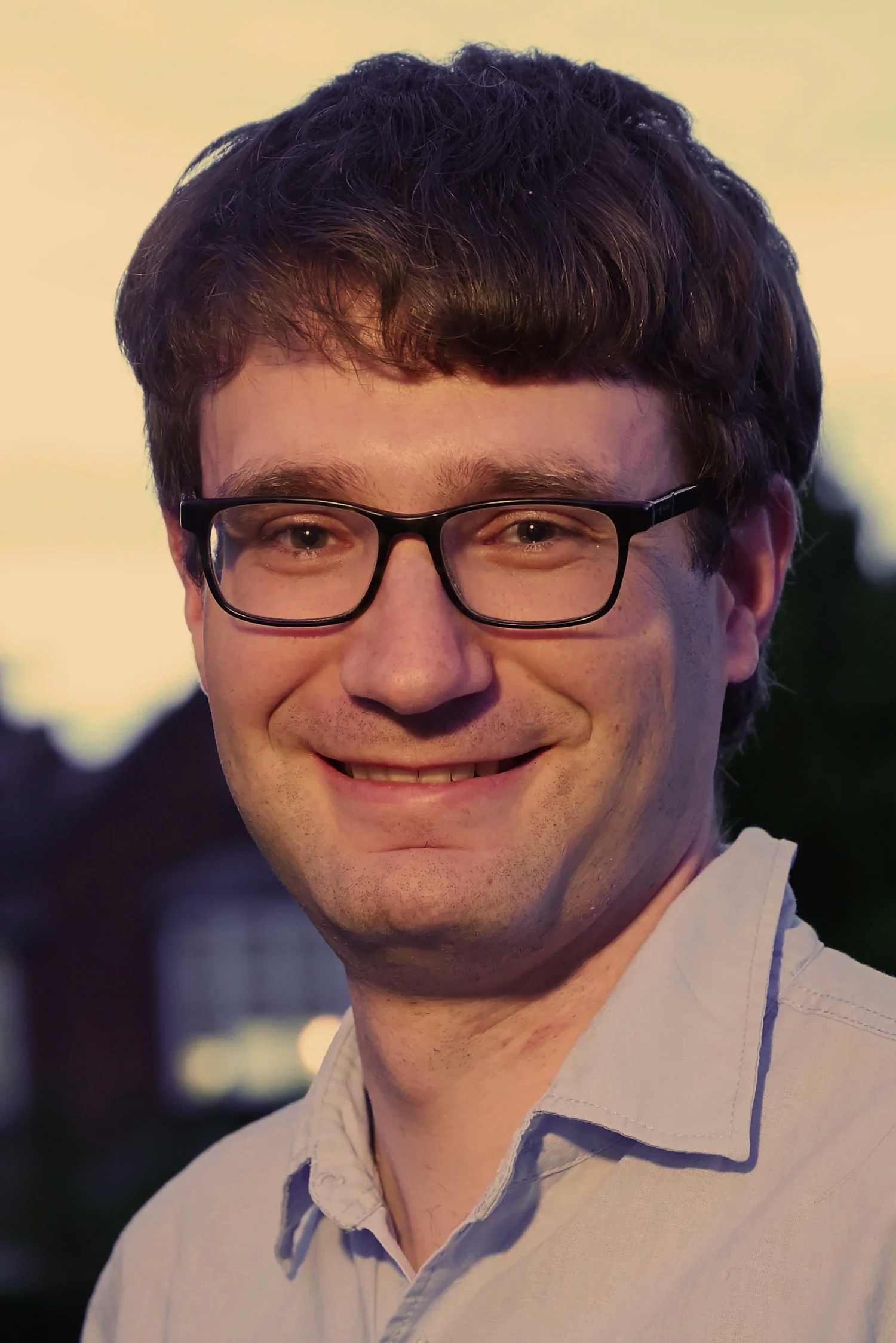 A young man with curly brown hair, wearing black glasses and a white shirt, smiling outdoors during sunset.