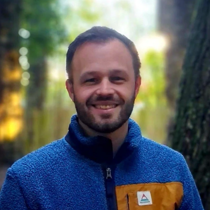 A smiling man with short hair and a beard, wearing a blue fleece jacket with a yellow pocket, standing outdoors in a forested area with trees and blurred green foliage in the background.