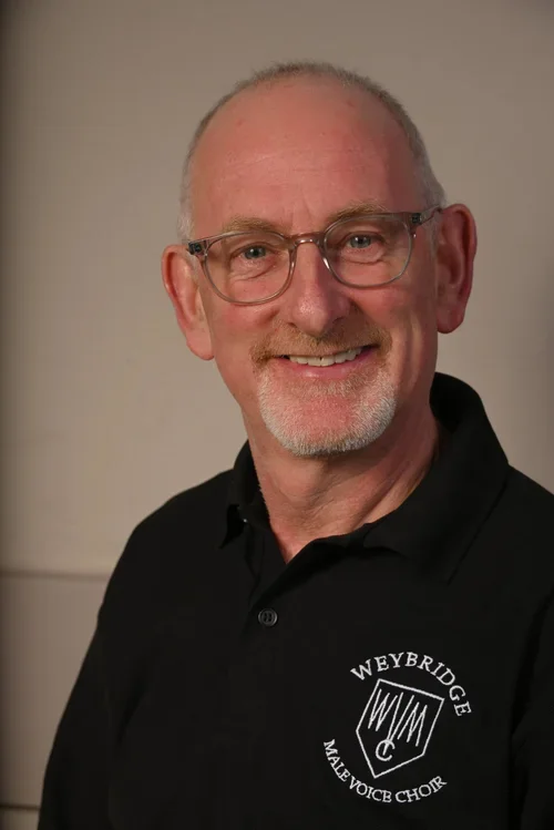 A smiling older man with glasses and a goatee, wearing a black polo shirt with a Weybridge Male Voice Choir logo, posing against a neutral background.