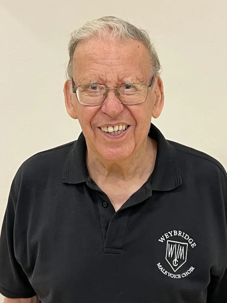 An elderly man with gray hair, wearing glasses, smiling and wearing a black polo shirt with the logo of Weybridge Male Voice Choir.
