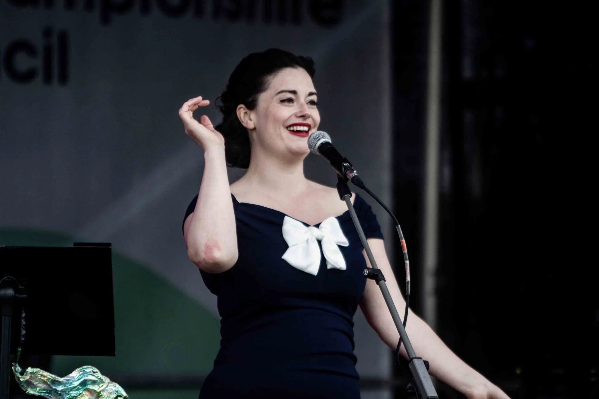 A woman with dark hair styled in waves, wearing a navy dress with a white bow on the neckline, smiling and speaking into a microphone on a stage.