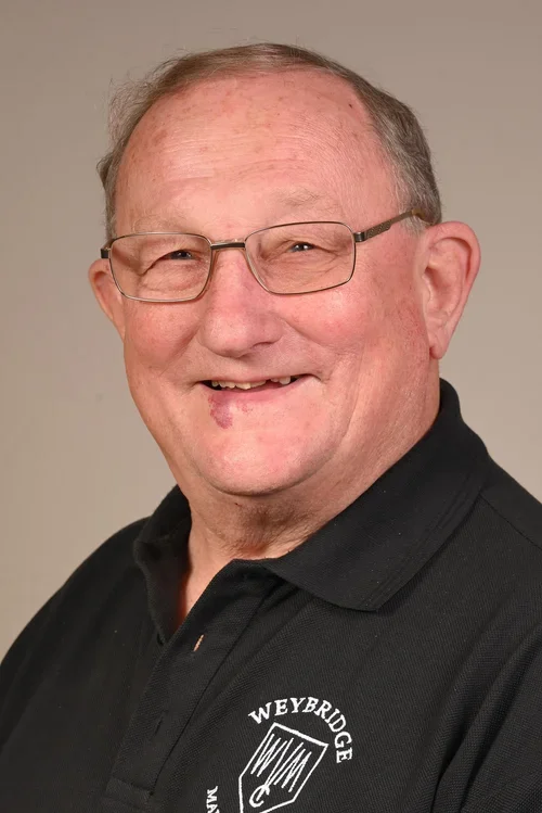 A smiling elderly man wearing glasses and a black polo shirt with a logo that says "Weybridge" and an emblem.