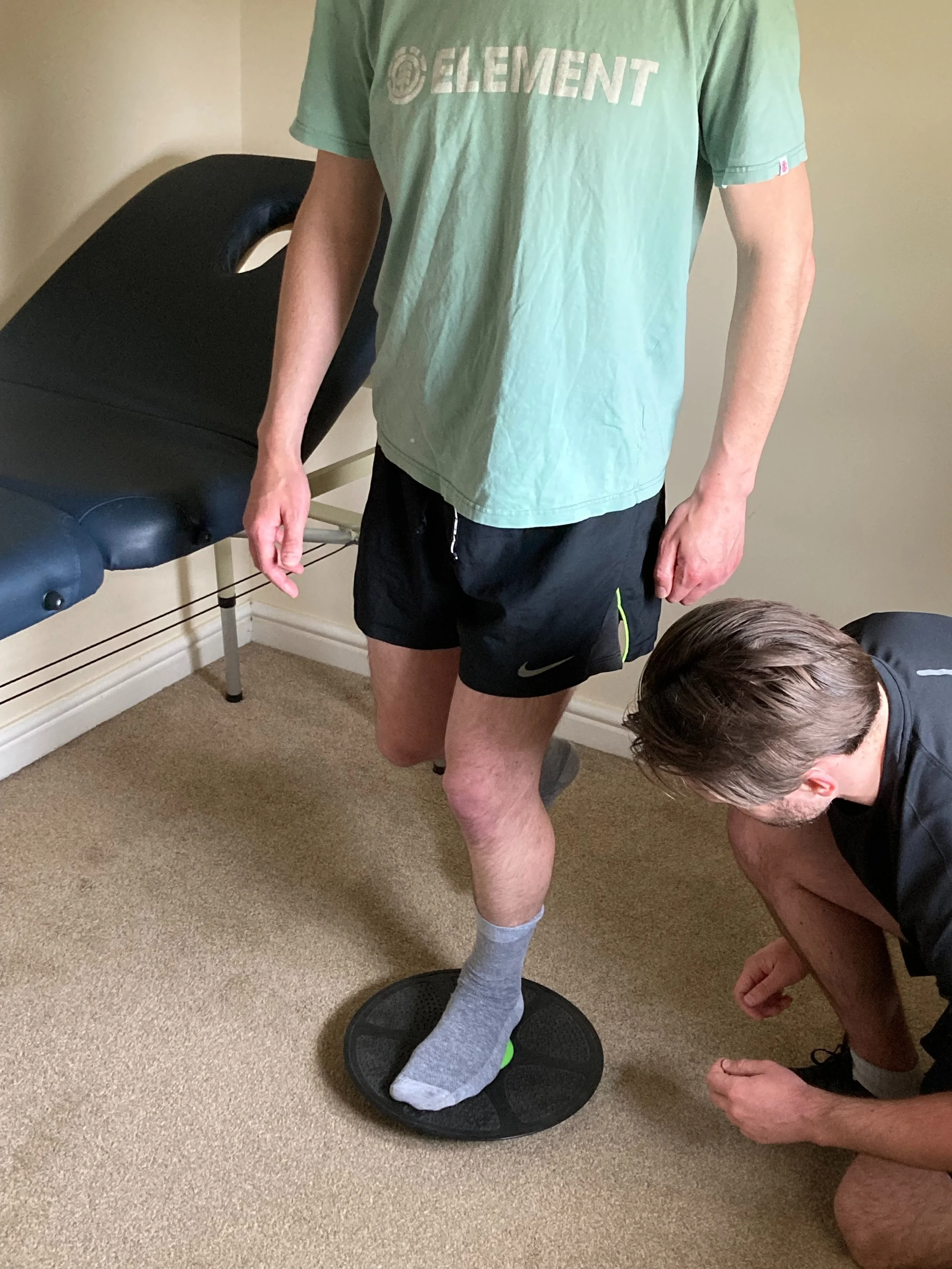 A person standing on a balance board with another person adjusting or inspecting his foot. The person standing is wearing a green T-shirt with the word 'ELEMENT' on it, black Nike shorts, and gray socks.