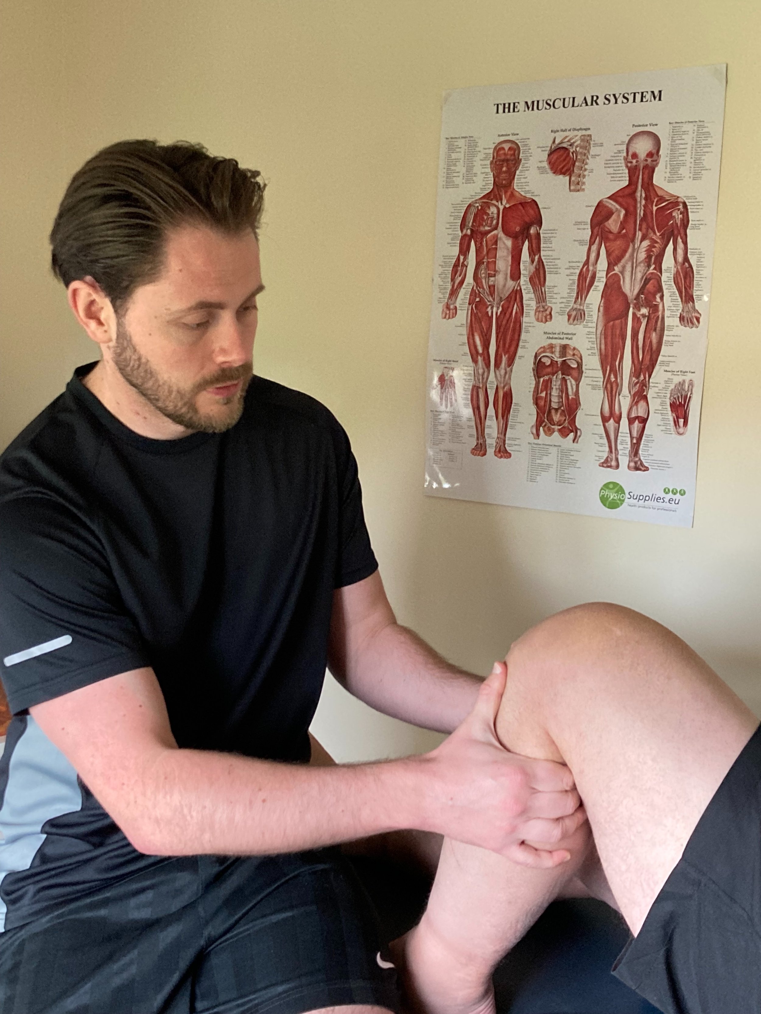 A man providing physical therapy to a patient's knee in a clinic room, with a detailed poster of the muscular system on the wall.