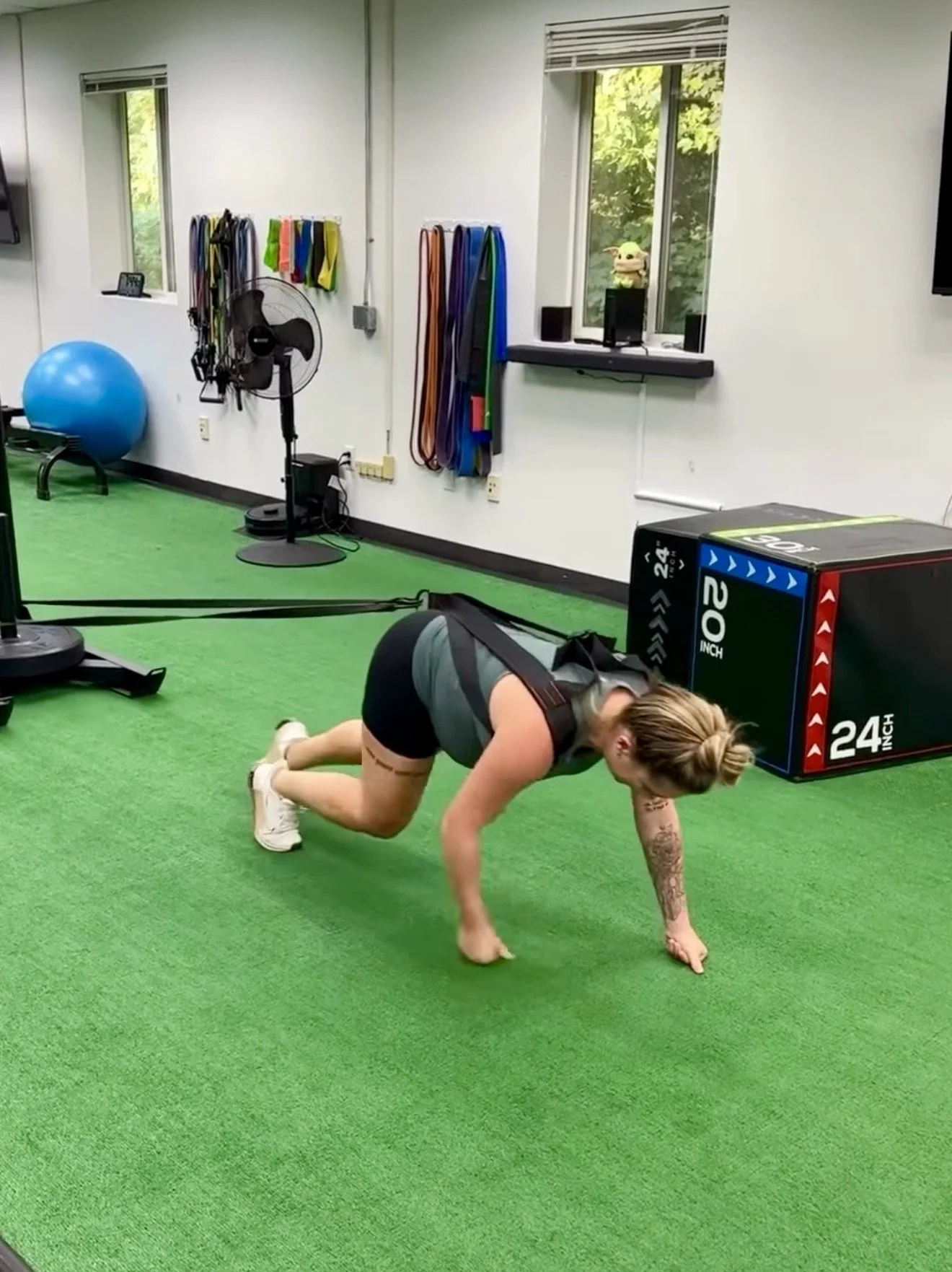 A pregnant woman doing a mountain climber exercise on a green turf in a gym with fitness equipment, resistance bands, a large exercise ball, and a window showing green foliage outside.