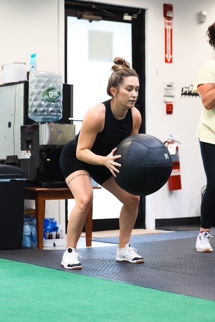 A woman in athletic attire doing a squat exercise with a large medicine ball in a gym setting.
