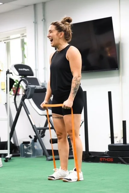 A woman in workout clothes using a resistance band at the gym, laughing and standing on a green turf floor.