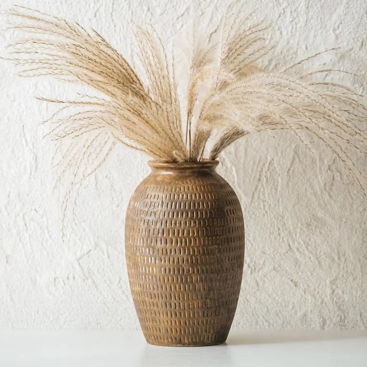 A textured brown ceramic vase filled with dried pampas grass, placed on a white surface against a plain textured wall.