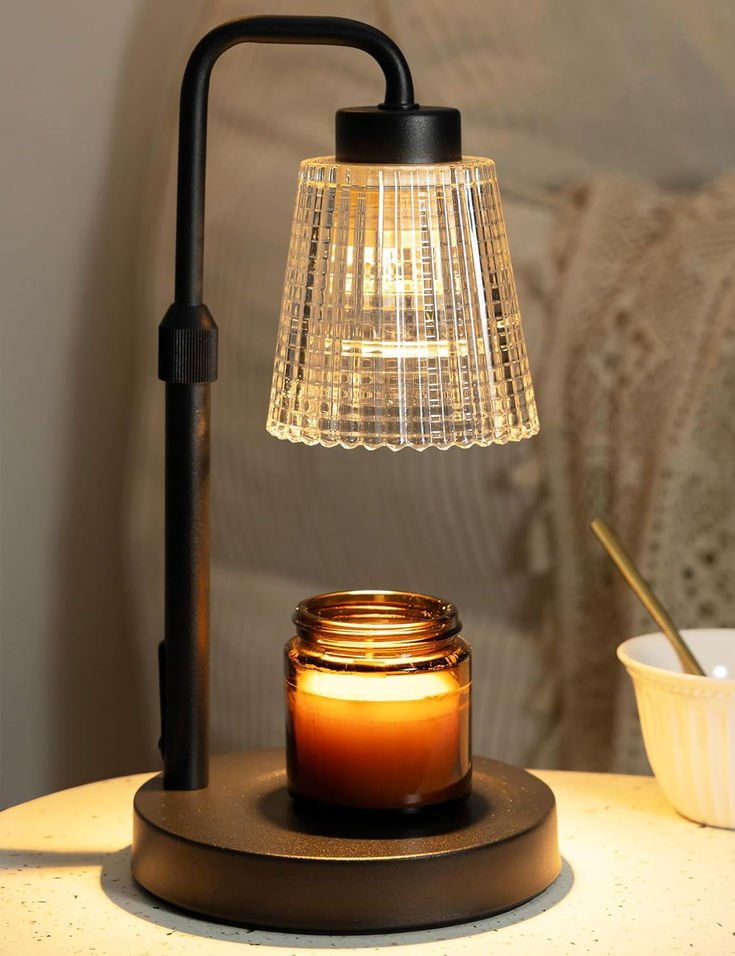 A lit candle in a brown glass jar is placed on a black circular base, illuminated by a vintage-style desk lamp with a glass shade. In the background, there is a white bowl with a spoon and a beige textured pillow or blanket.