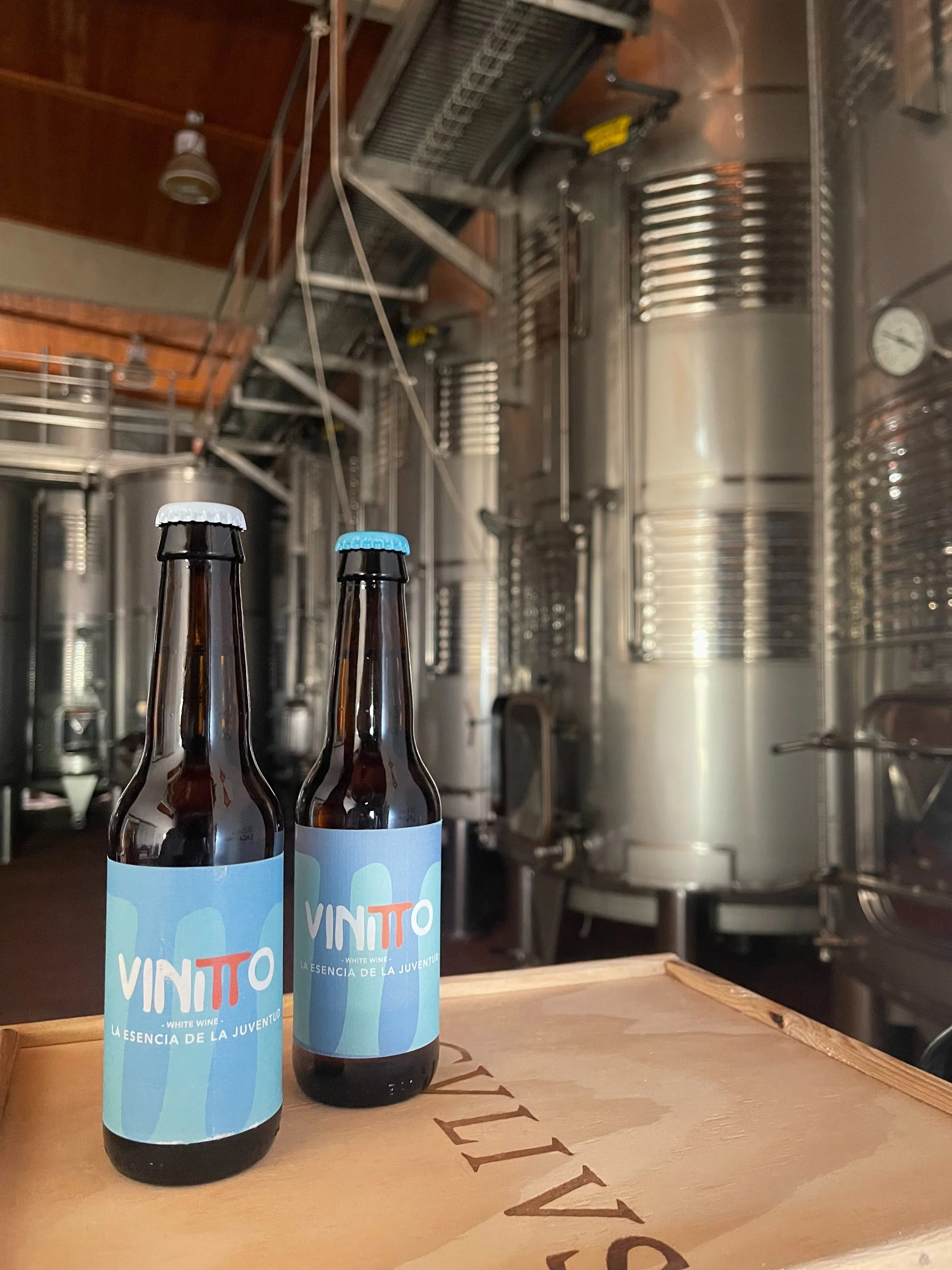 Dos botellas de vinito blanco con etiquetas azules y blancas en una mesa de madera, fondo de una bodega con tanques de acero inoxidable.