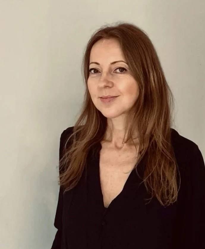 A woman with long, reddish-brown hair and light skin, wearing a black top, standing against a plain, light-colored wall.