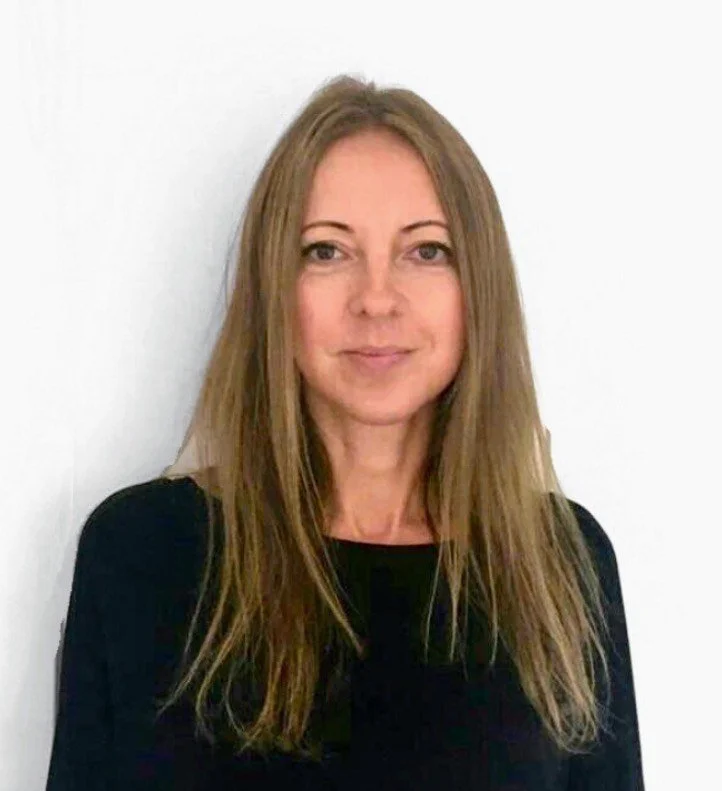 A woman with long, light brown hair wearing a black top, standing against a white background.