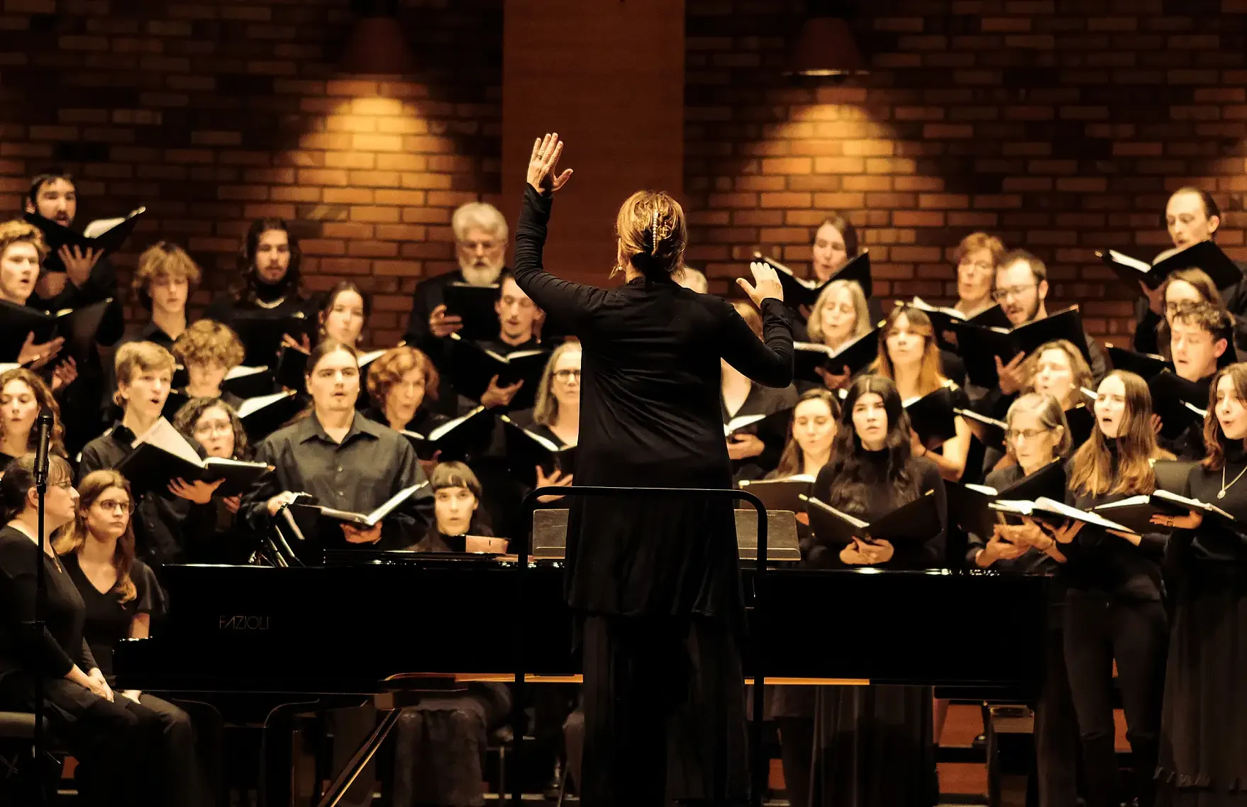 Conductor leading a choir of singers in a performance inside a theater with a brick wall background.