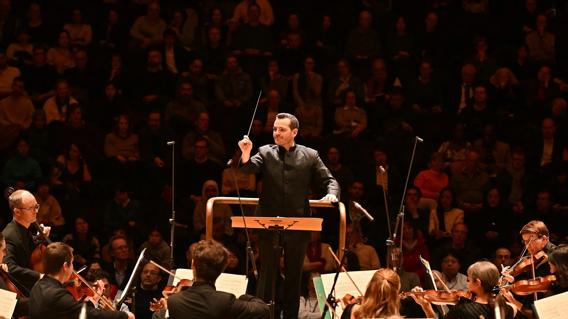 A conductor leads an orchestra during a performance, standing on a podium with a music stand, with musicians playing string instruments and an audience watching in the background.