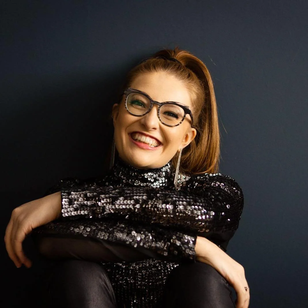 A young woman with red hair, glasses, and a big smile, wearing a black sequined top, sitting against a dark background.