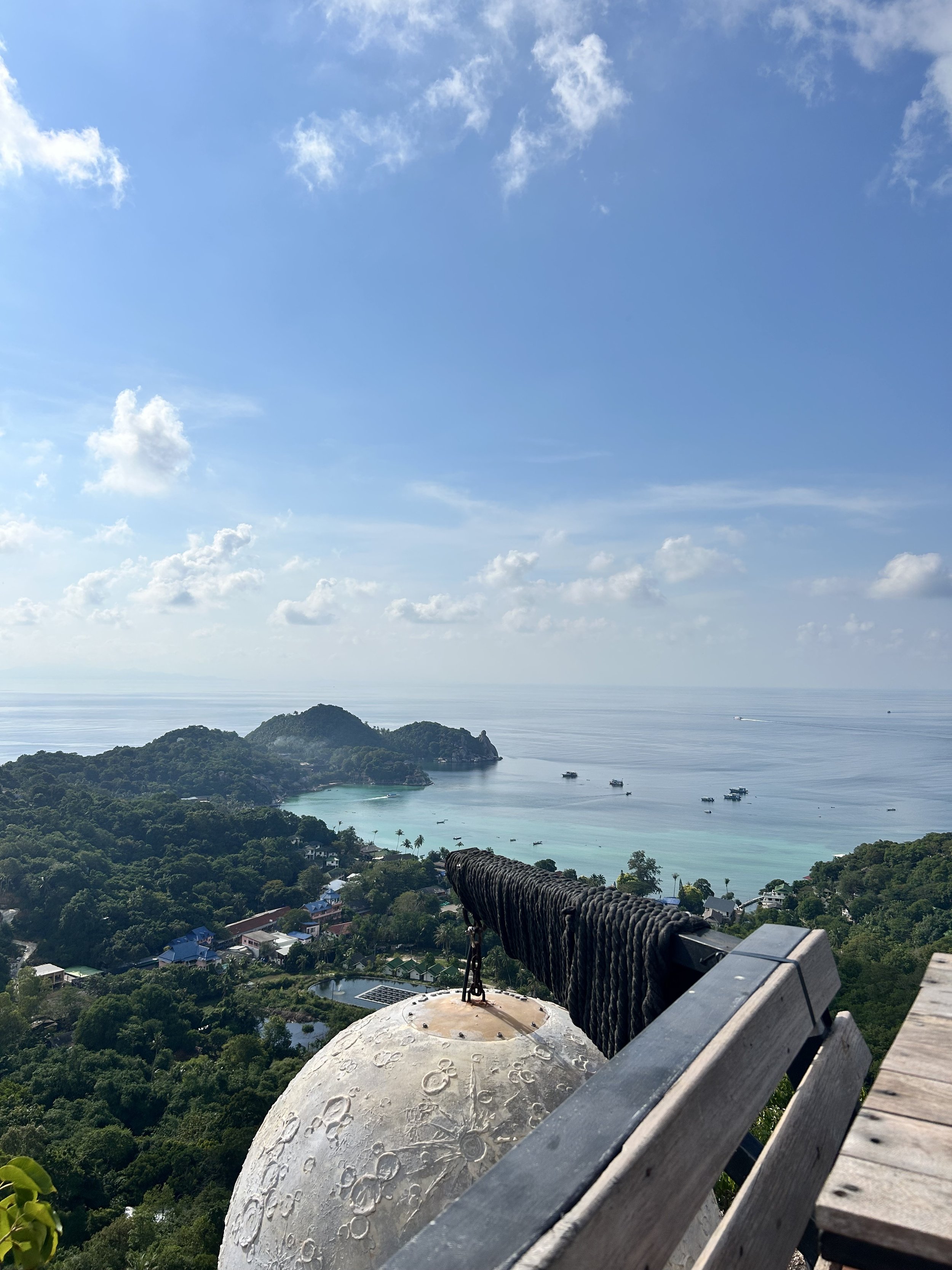 View from a high vantage point showing a tropical coastal landscape with lush green trees, a bay with turquoise water, several boats, and distant islands under a partly cloudy sky.