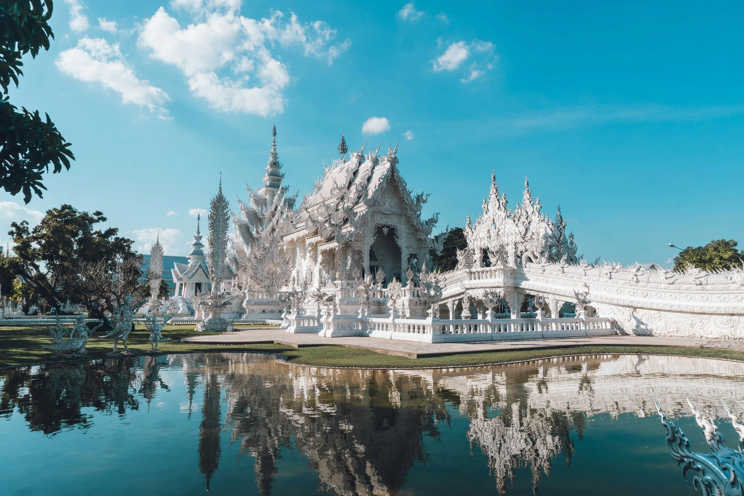 Bright white temple with intricate details, reflected in water, under a blue sky with scattered clouds.