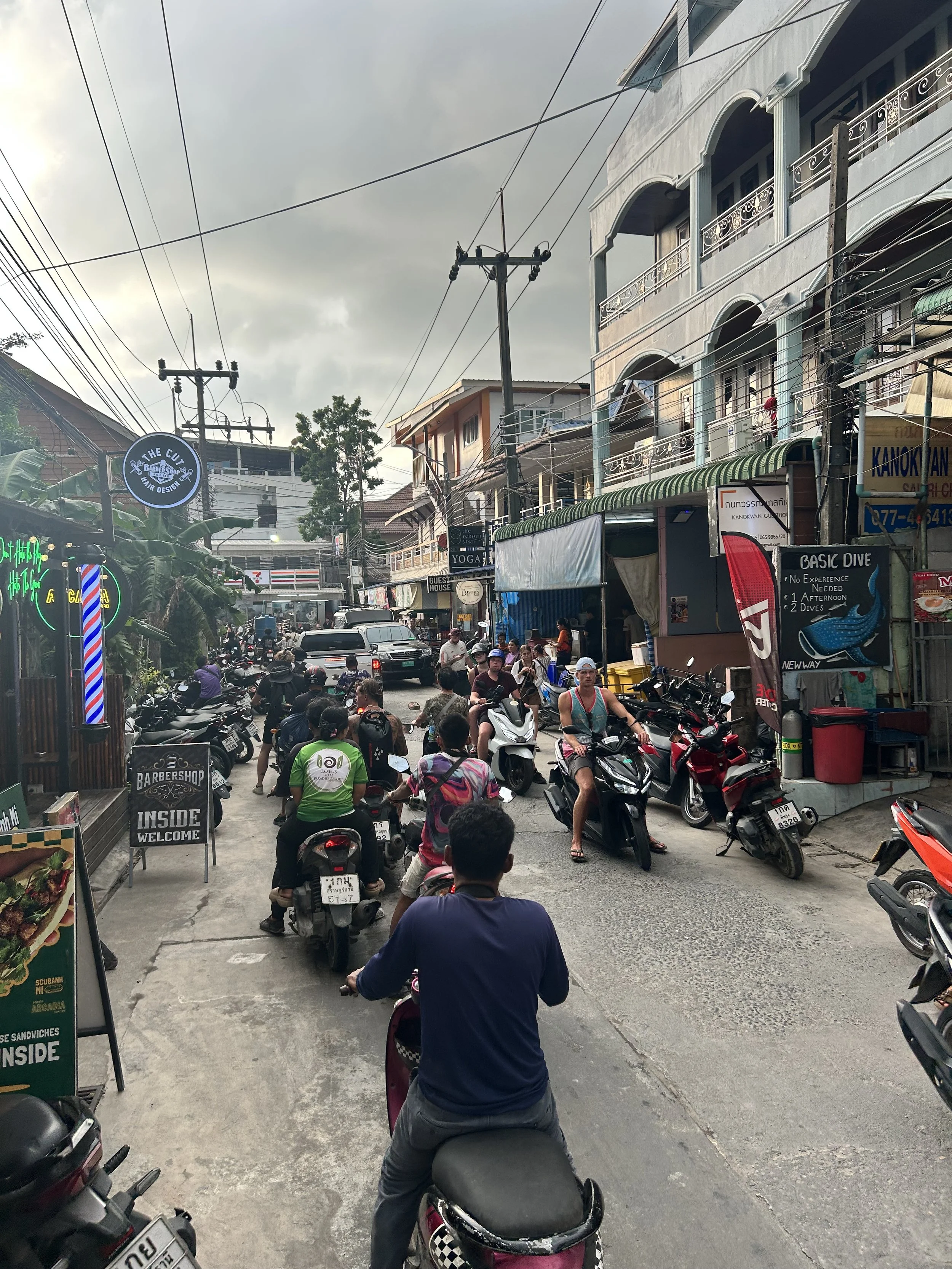 A bustling street scene with many motorbikes and scooters, people walking and waiting, shops, signs, and overhead power lines in an urban area.