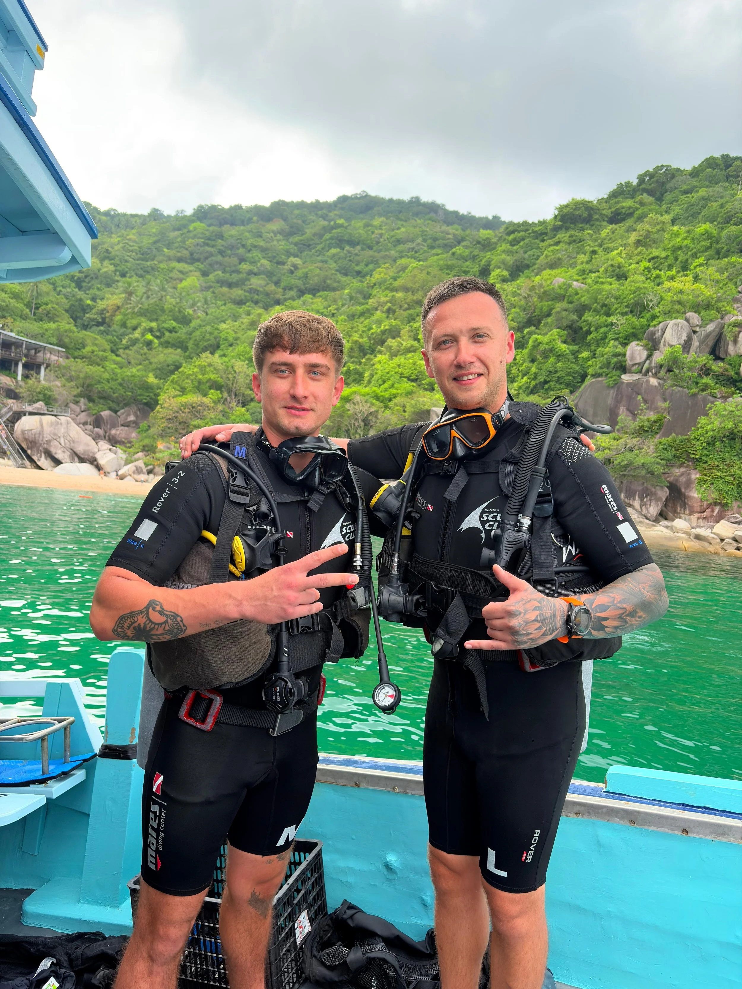 Two men in scuba diving gear standing on a boat with a lush, green hillside and rocky landscape in the background.
