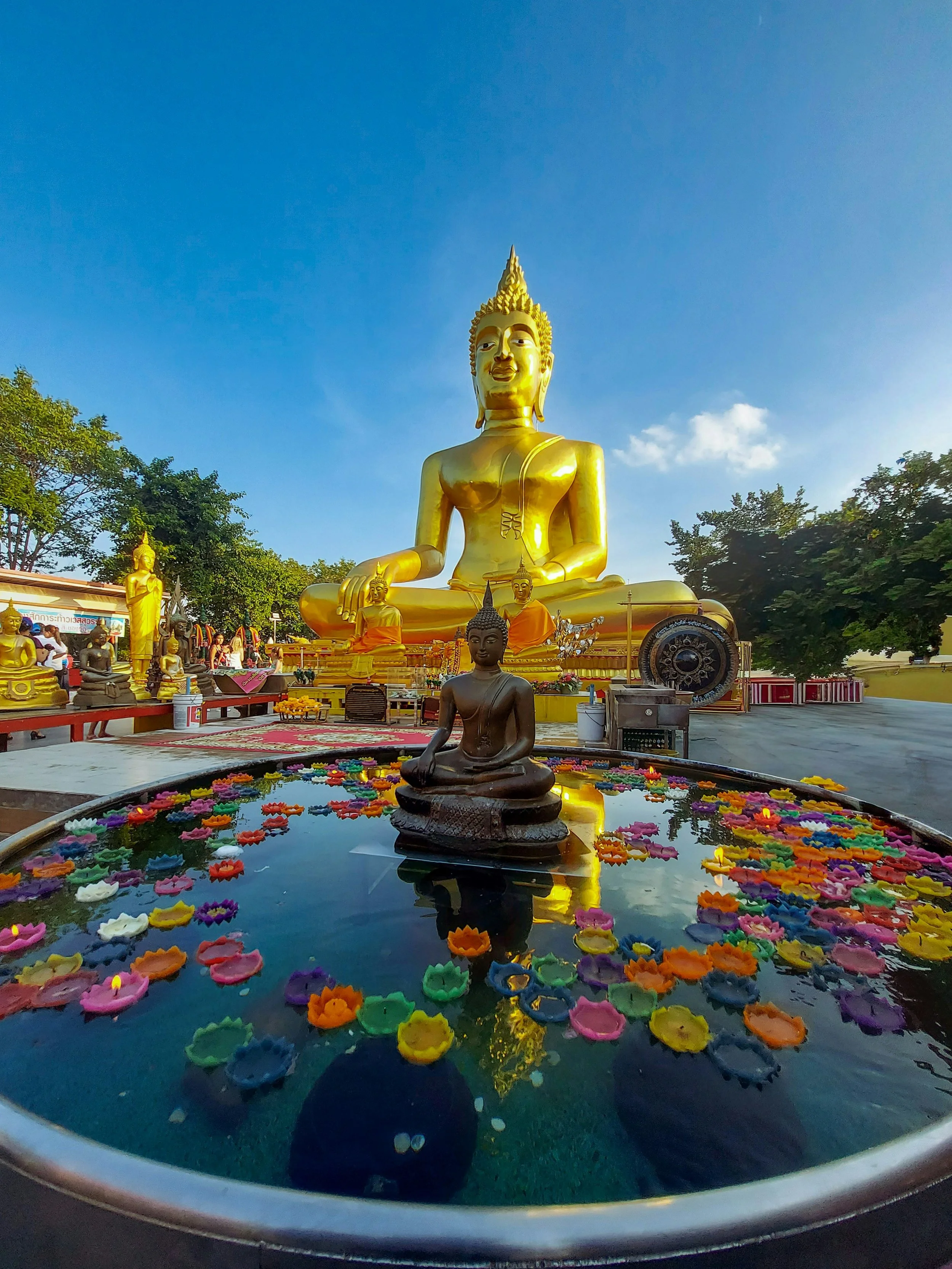 Golden Buddha statue seated in meditation pose, surrounded by smaller statues, with a small shrine and floating candles on a water basin in the foreground, under a blue sky.