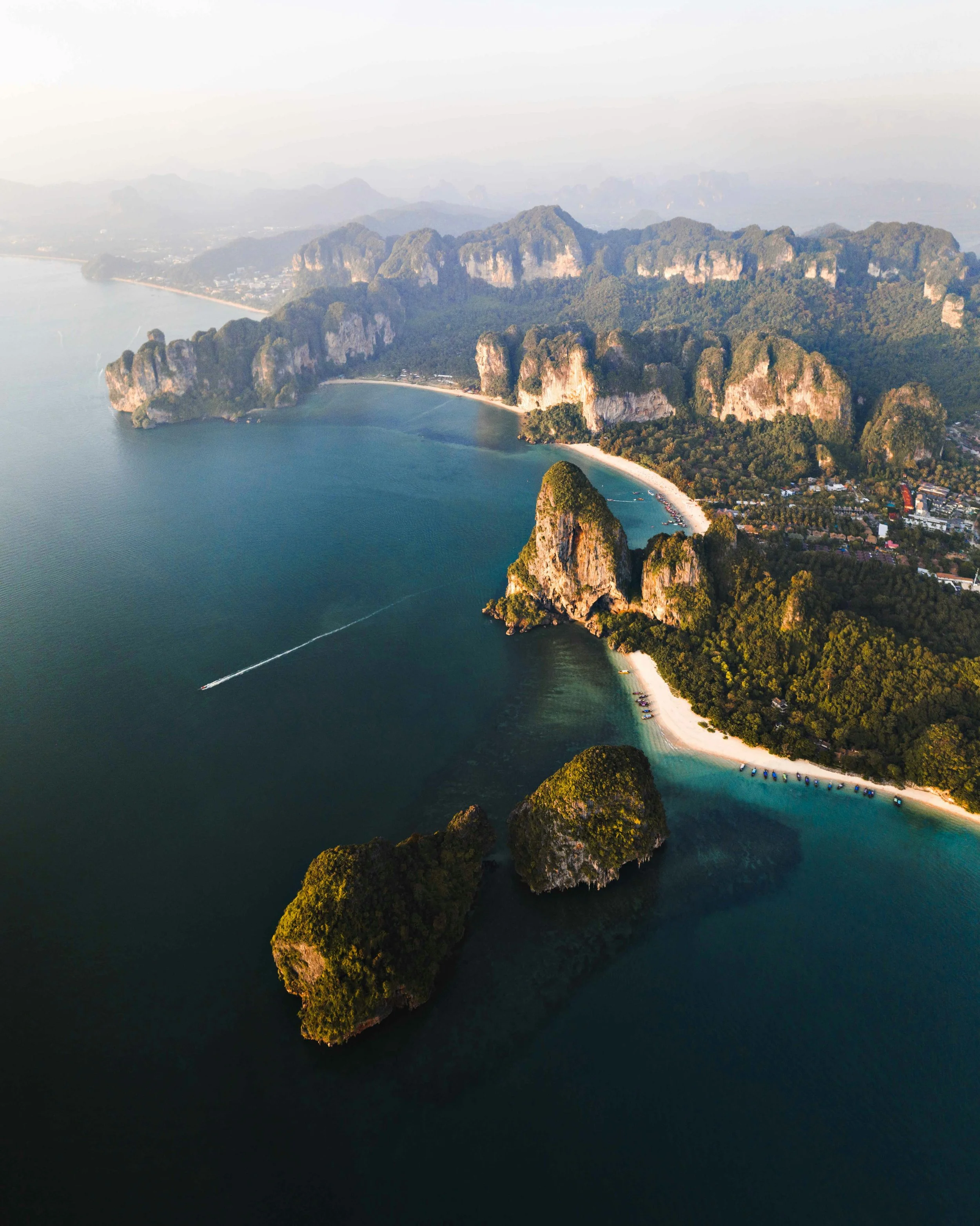Aerial view of limestone islands and beaches along a tropical coast, with boat trails in the water and lush green foliage.