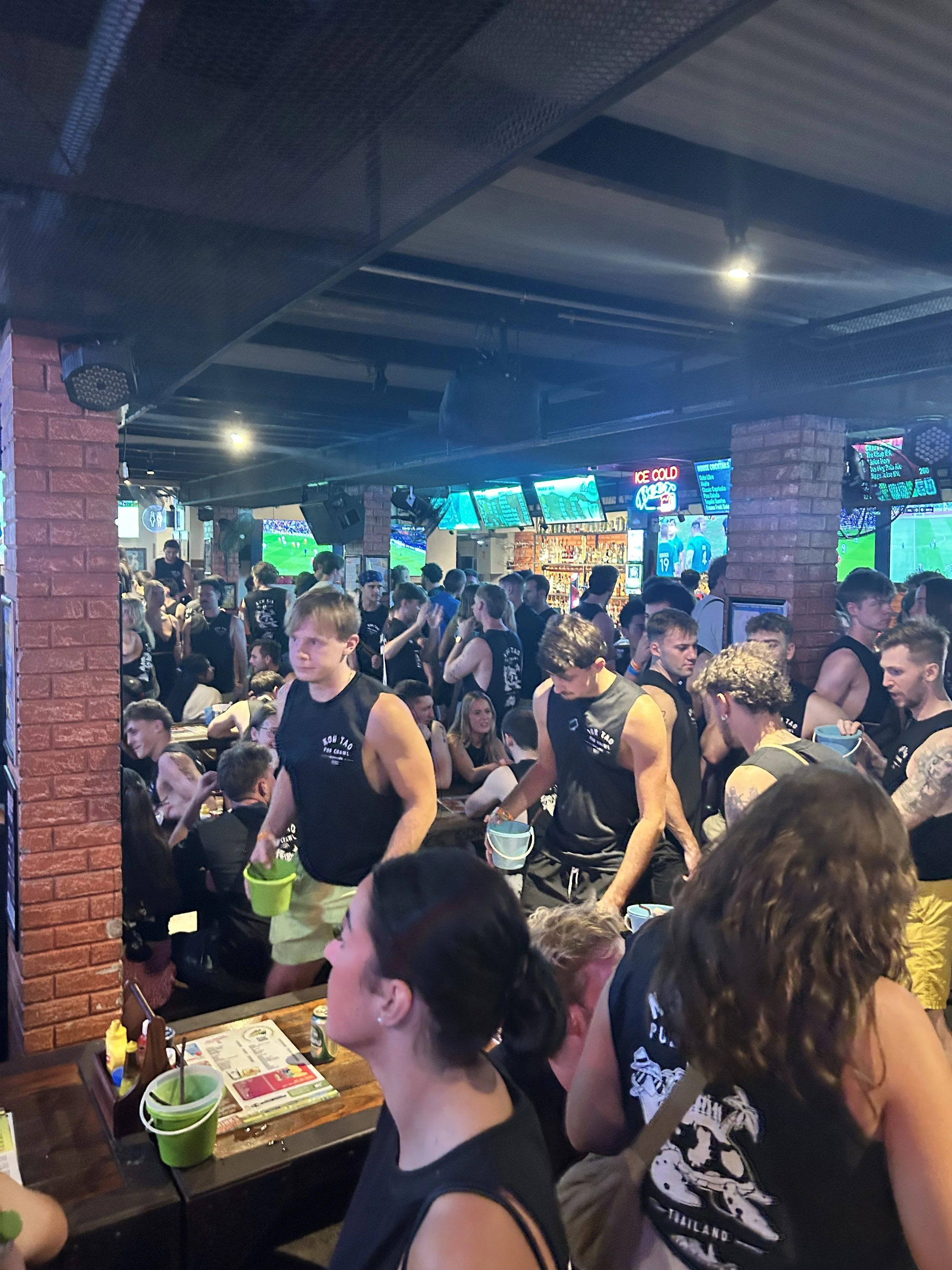 Crowded sports bar with people watching multiple television screens showing a sporting event, some seated and some standing, with dim lighting and exposed brick walls.