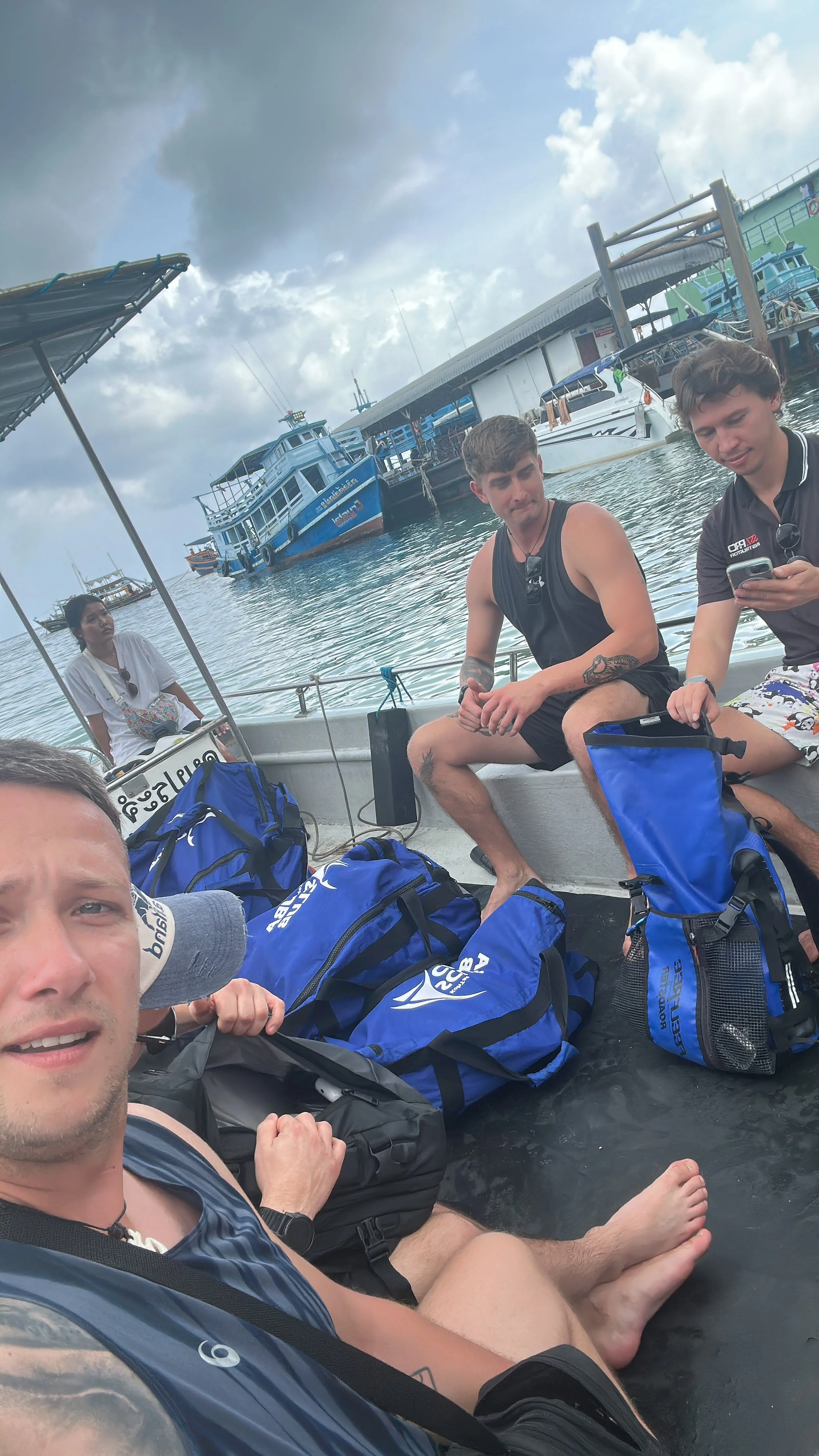 Group of young people sitting on a boat near a dock, with blue backpacks, boats, and a cloudy sky in the background.