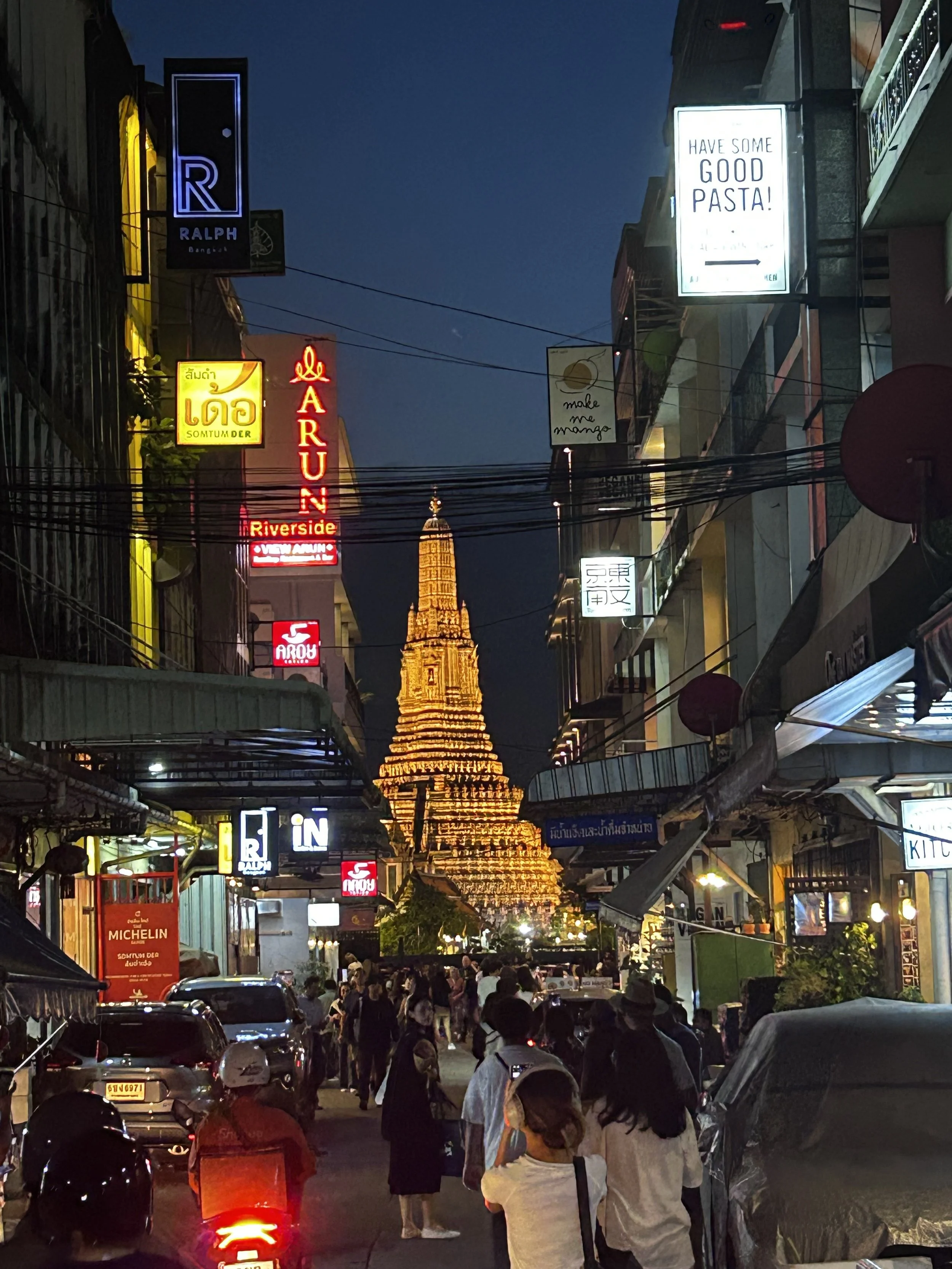 Night scene of a busy street with illuminated signs in a foreign language, crowded with people and vehicles, leading to a lit-up golden temple or pagoda in the background.