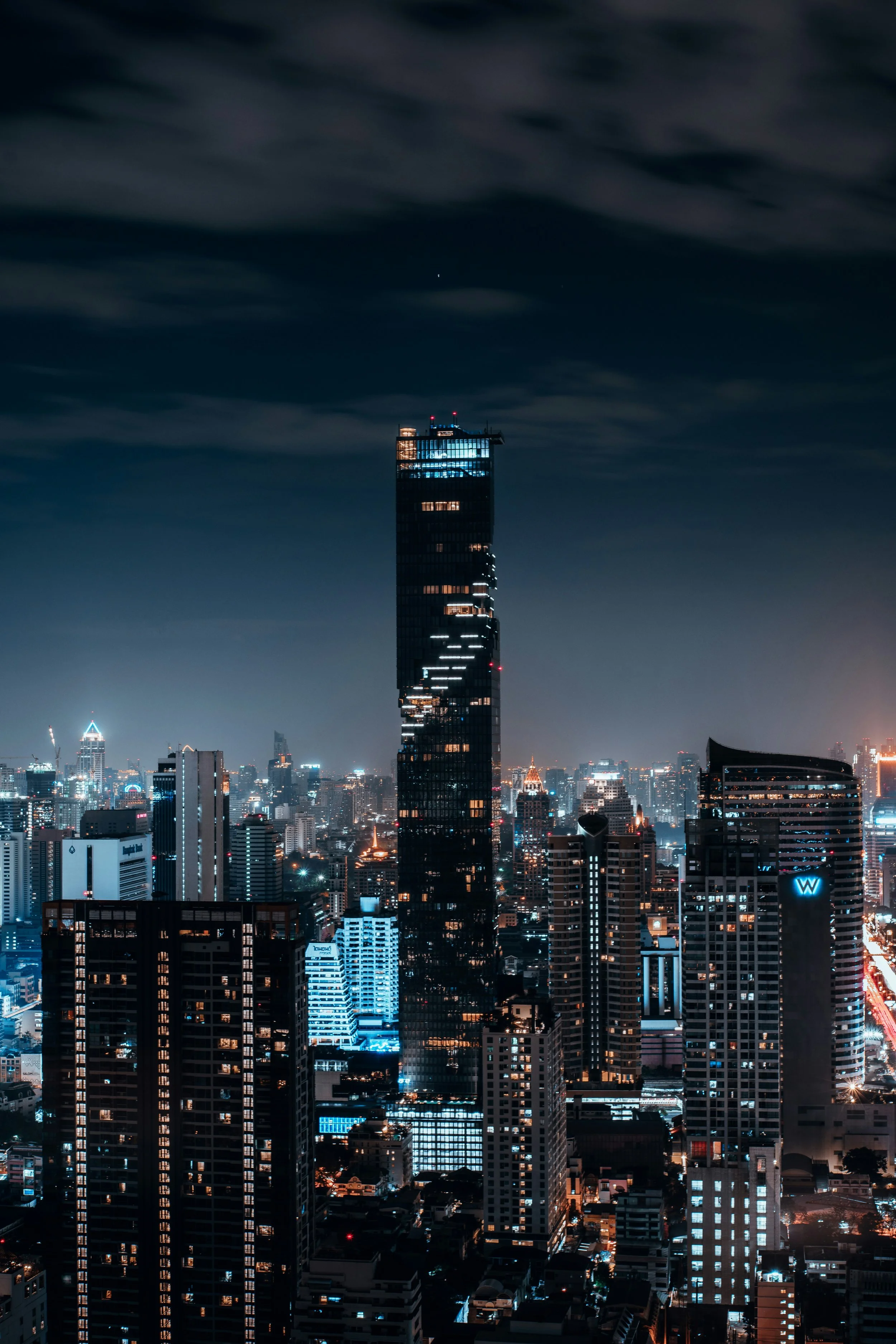 Nighttime cityscape with a tall illuminated skyscraper in the center, surrounded by other high-rise buildings and city lights.