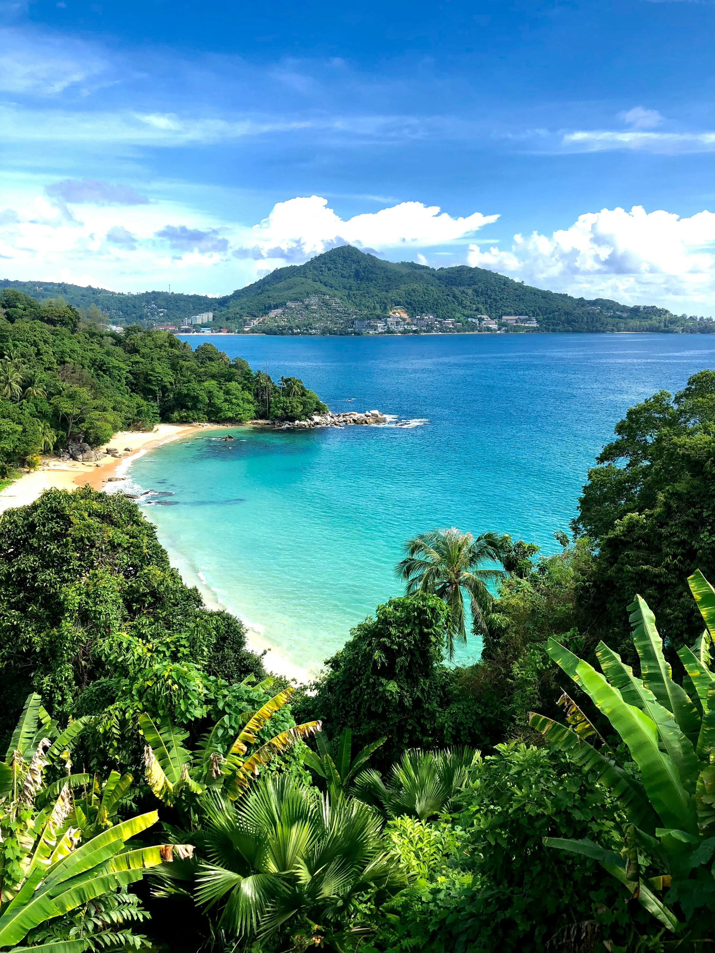 Tropical beach with turquoise water, sandy shoreline, lush green vegetation, and a mountain in the background under a partly cloudy sky.