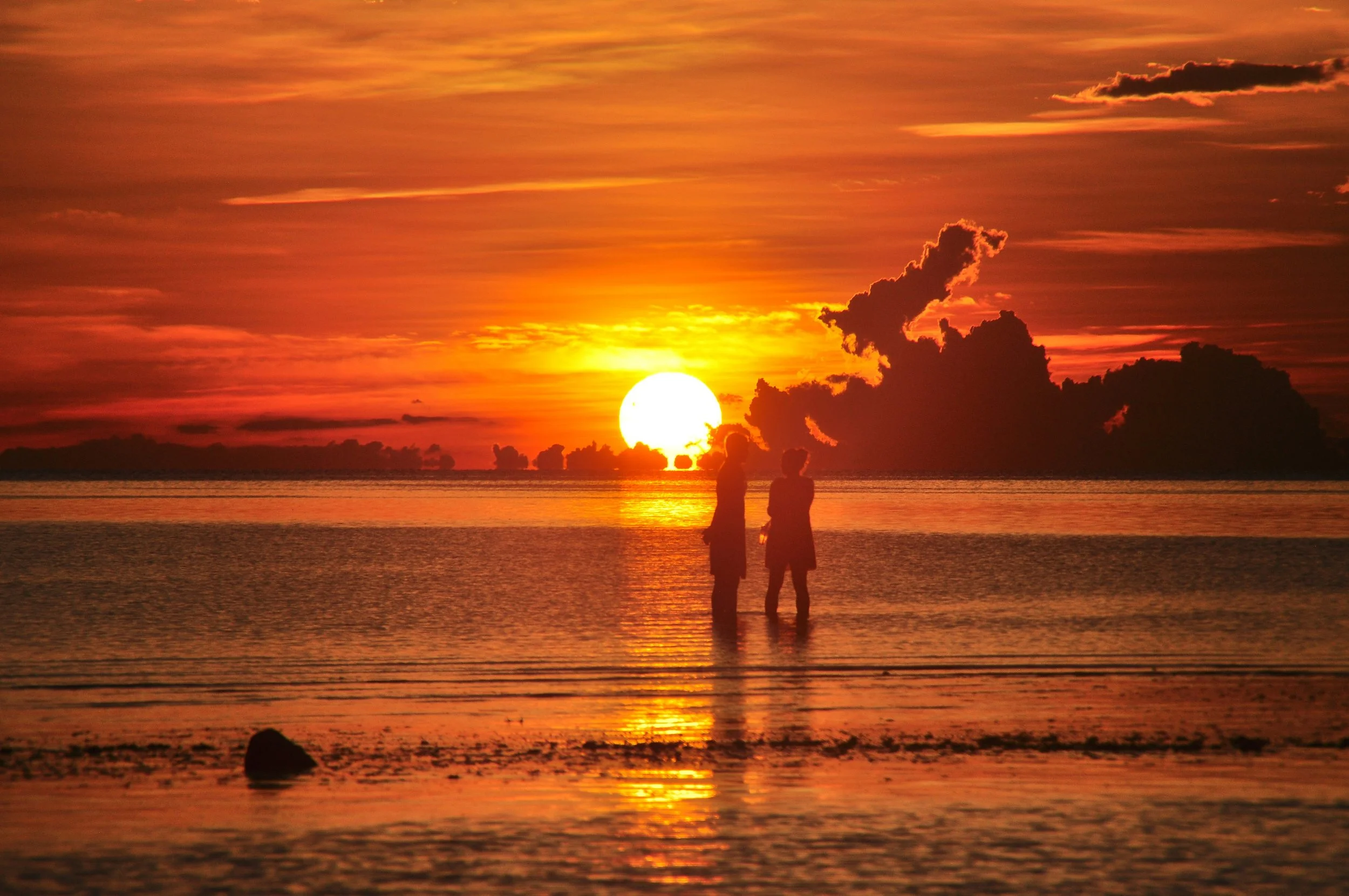 Silhouettes of two people standing in water during a sunset with orange and yellow sky, clouds, and reflections on the water.
