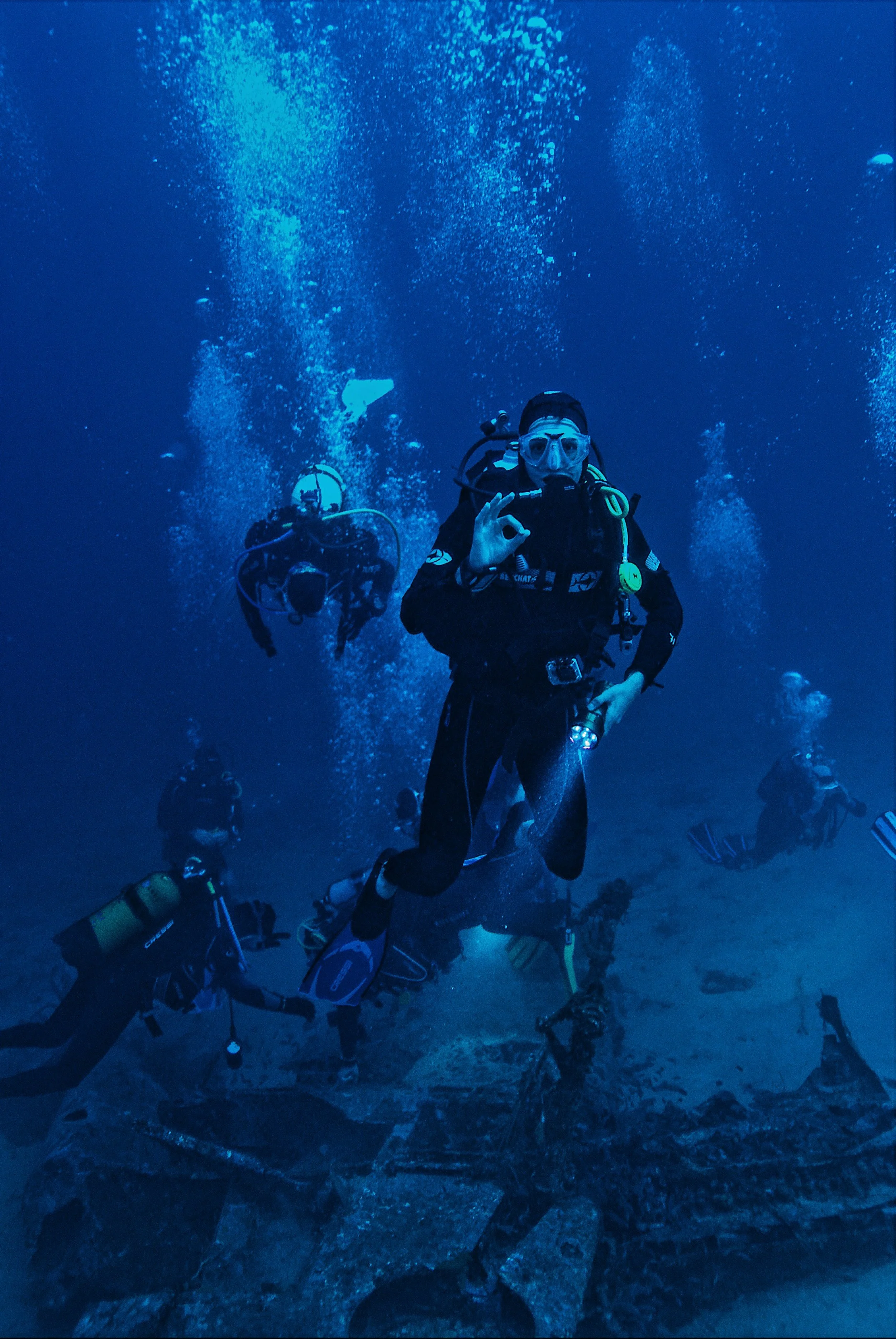 Scuba divers underwater, pose and communicate with hand signals, surrounded by bubbles and rocks.