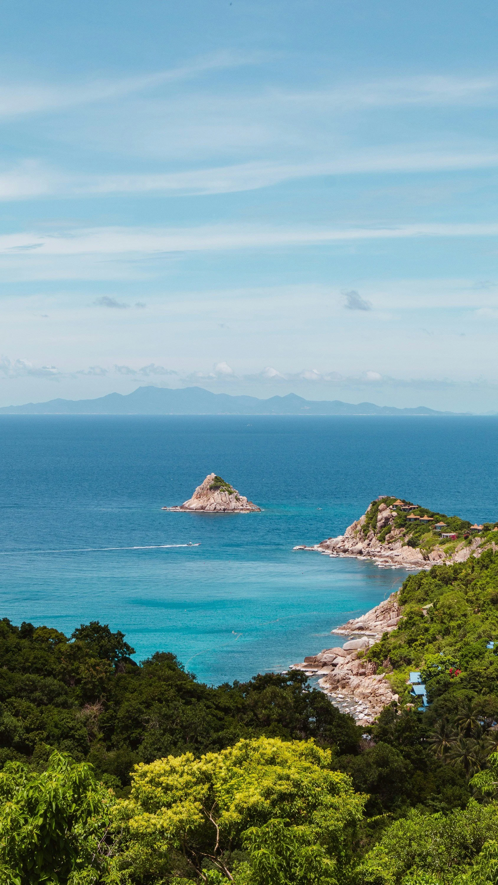 Scenic view of a rocky coastline with greenery, calm blue ocean, and a small island in the distance under a partly cloudy sky.