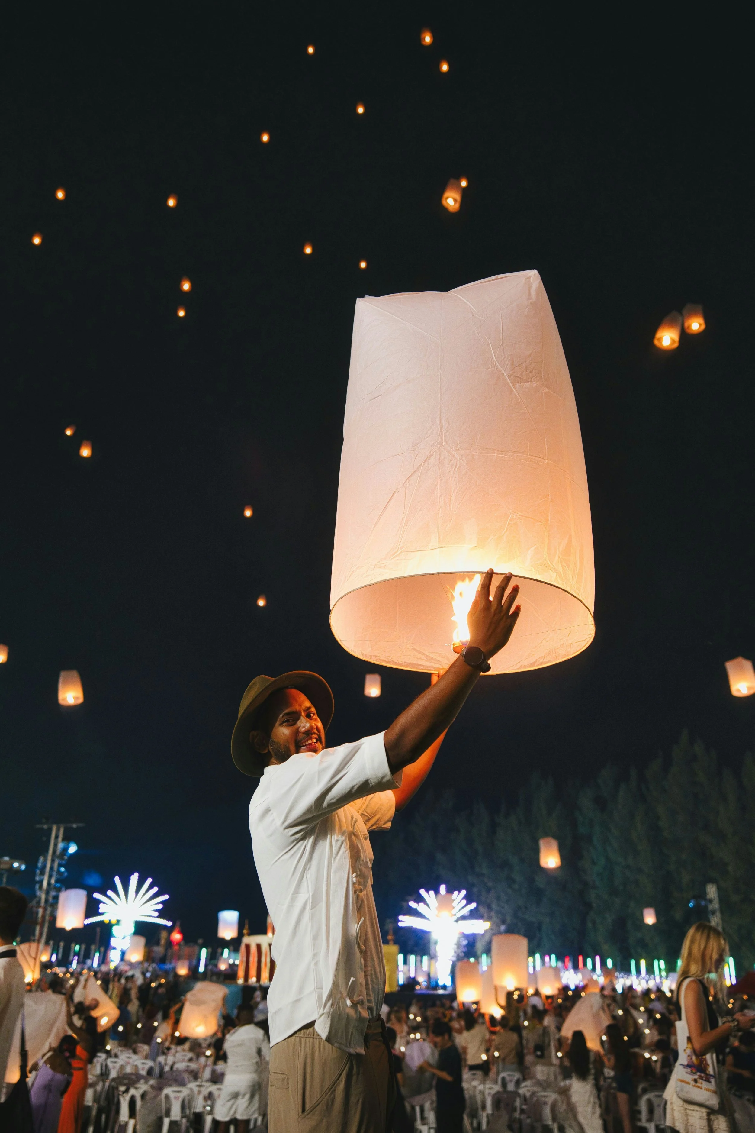 A man at night releasing a sky lantern during a festival, surrounded by many other paper lanterns floating in the sky and colorful lights in the background.