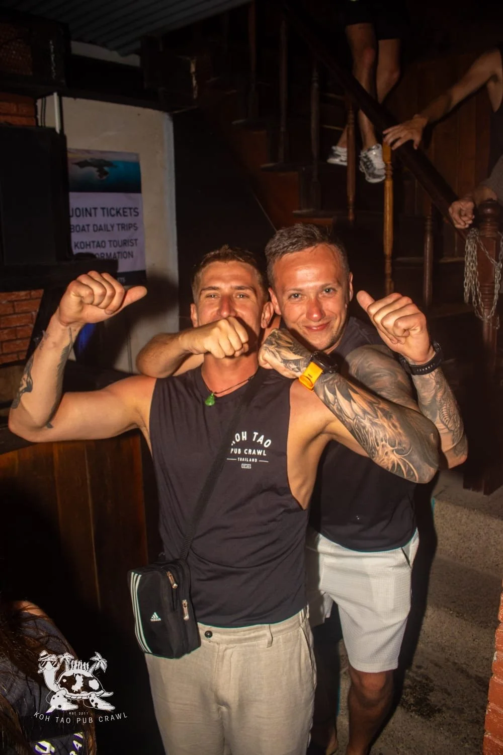 Two men are posing with their arms flexed, smiling and showing strength, inside a dimly lit bar or pub. One has a tattooed arm and is wearing a black tank top, while the other has a tattooed arm and is wearing a black T-shirt and light-colored shorts.