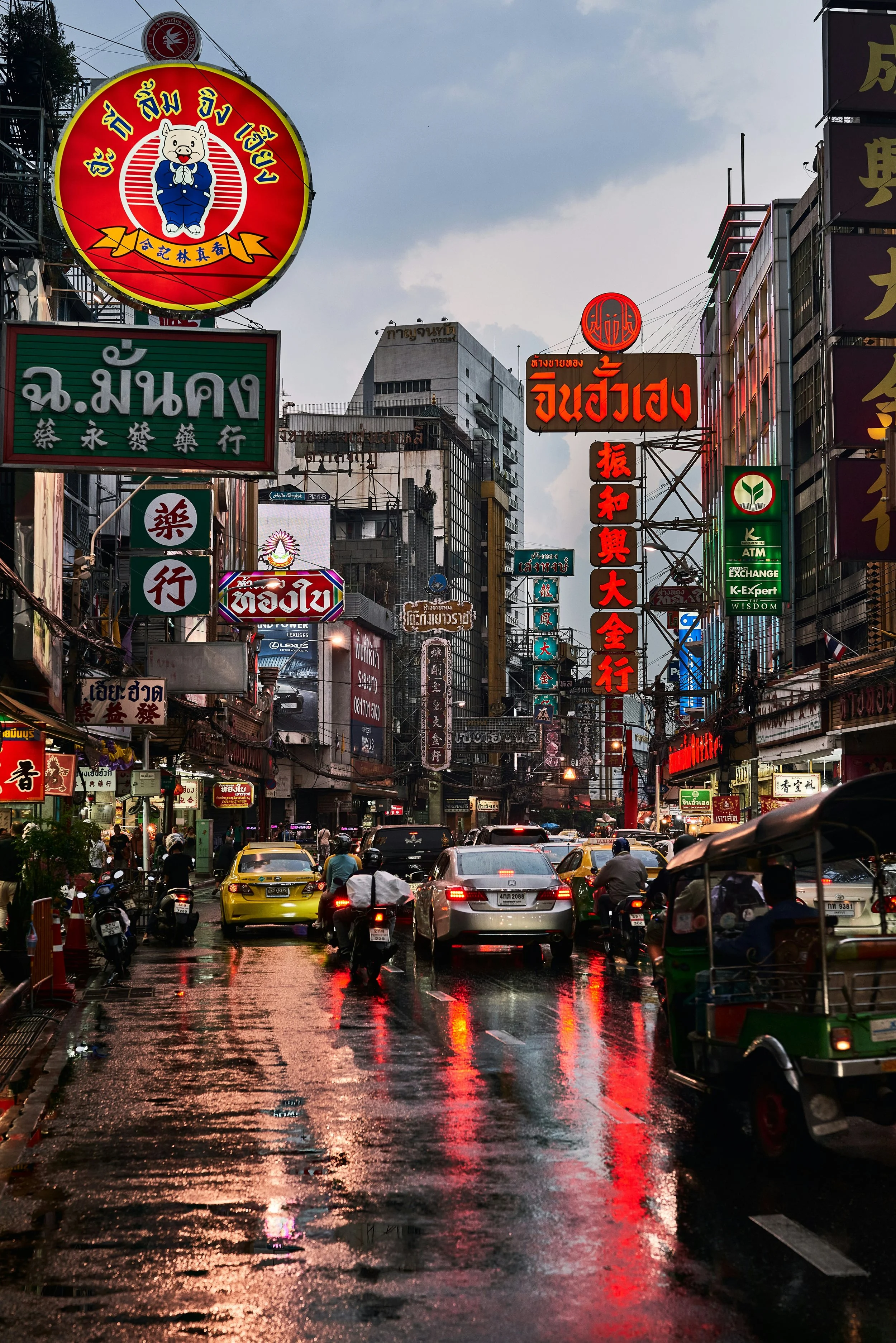 A busy city street at dusk with wet pavement reflecting colorful neon signs in Thai and Chinese characters, with cars and motorcycles moving through.