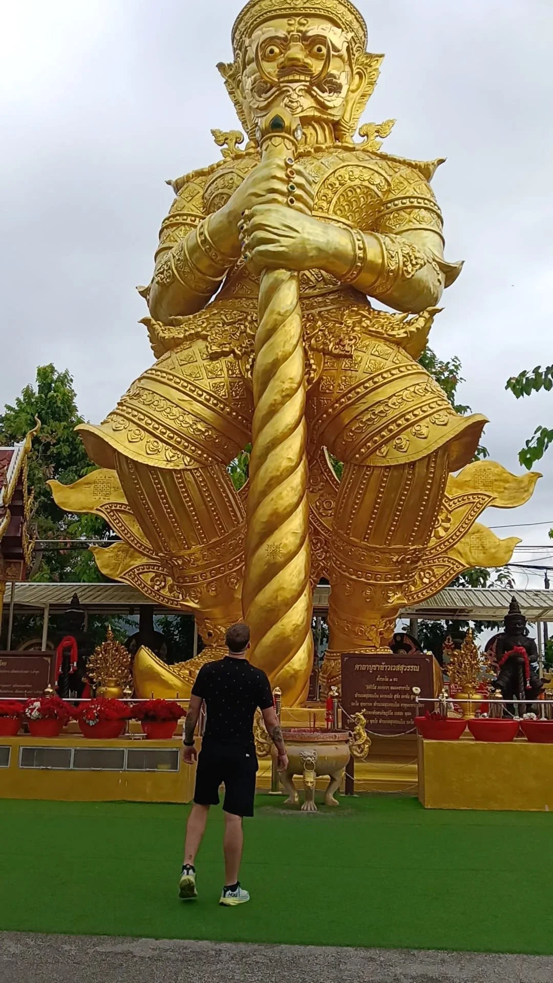 A large golden statue of a figure with armor, holding a large spiral staff, in a temple courtyard with a man in black clothing standing in front of it.