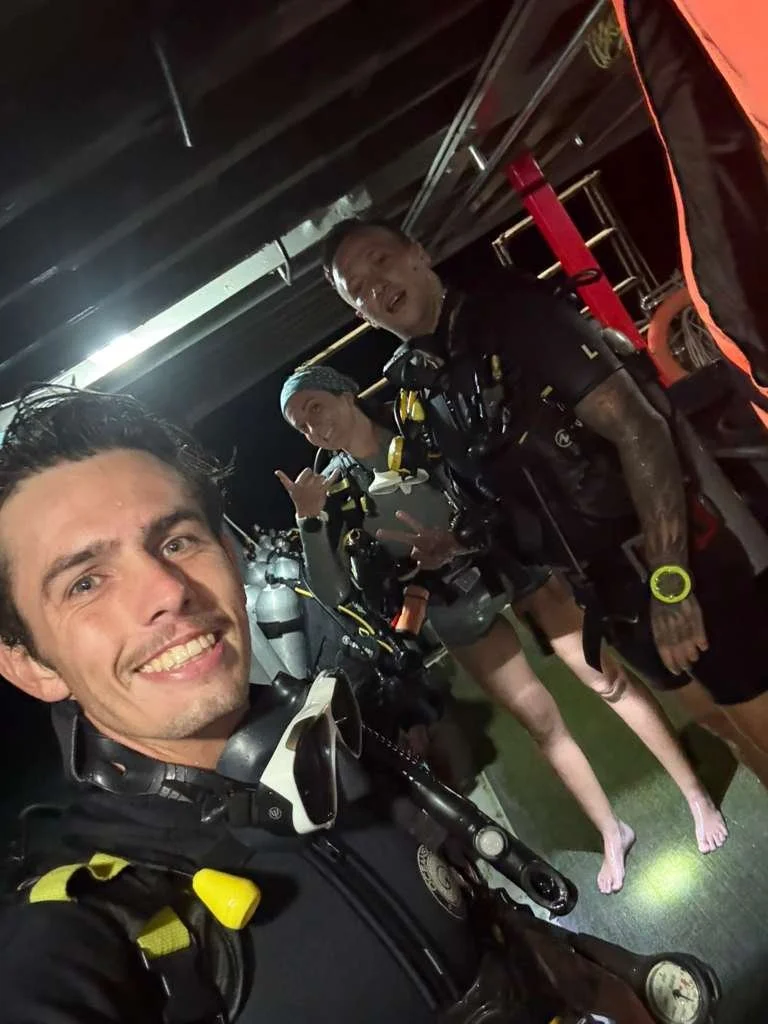 Three scuba divers in wet suits and diving gear smiling and posing for a selfie inside a boat or dock area, with a dark background and some equipment visible.