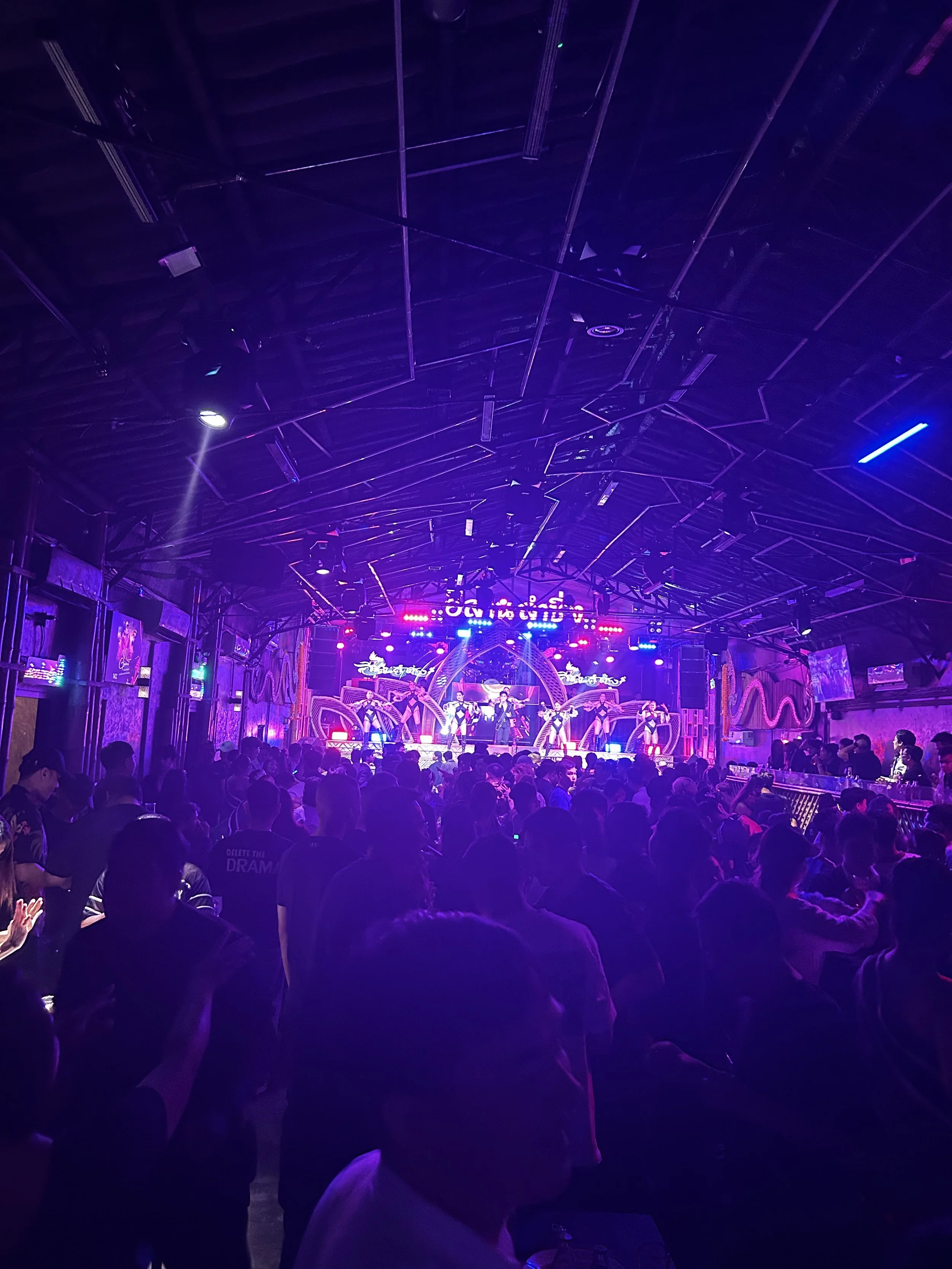 Concert or performance with a stage, colorful lighting, and a crowd of people standing and sitting in front, watching performers on stage.