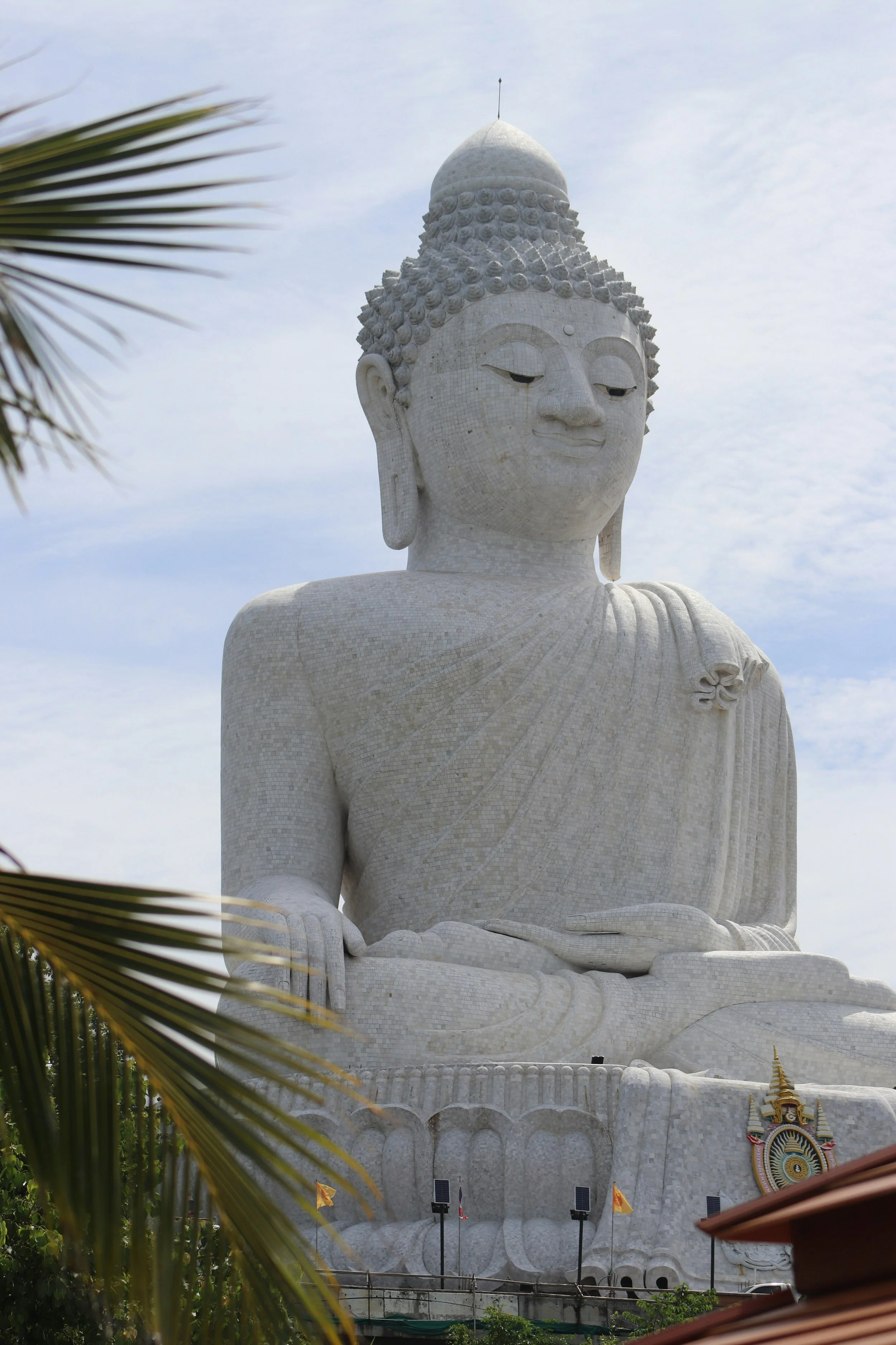 Large white Buddha statue with a serene expression, seated in a meditative posture, with palm leaves in the foreground and a cloudy sky in the background.