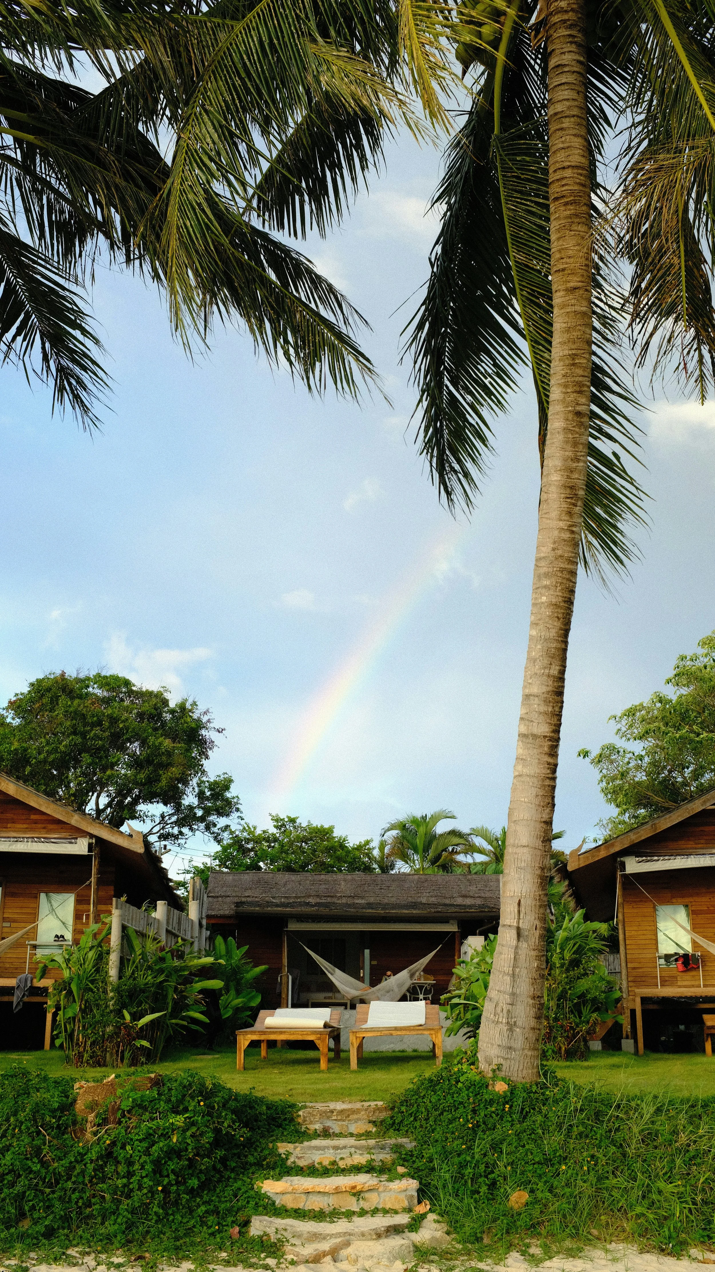 A tropical backyard with two wooden cabins, hammocks, lounge chairs, lush greenery, palm trees, and a rainbow in a partly cloudy sky.