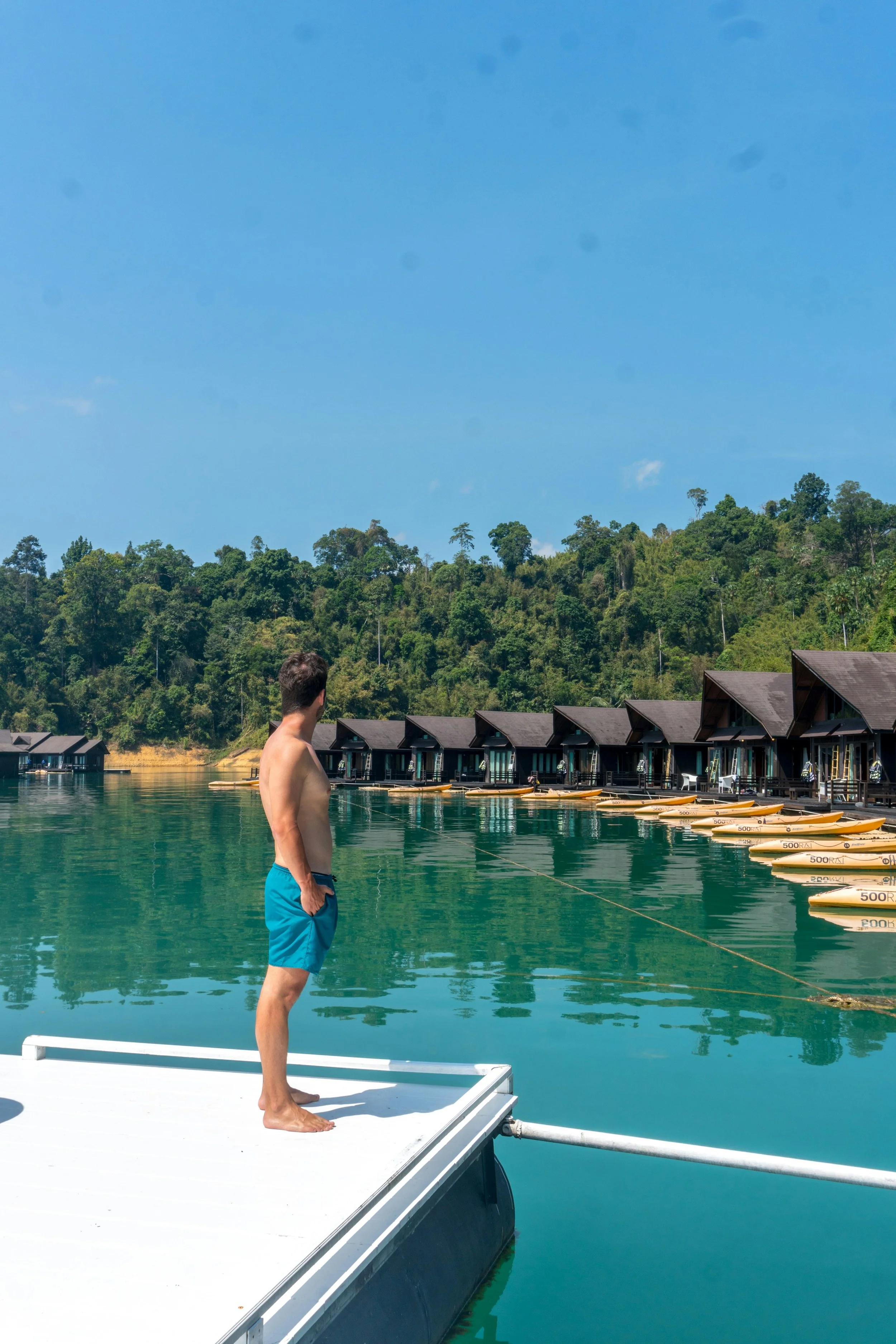 A man in blue shorts stands on a dock, looking at a row of overwater bungalows and kayaks lined up along a calm, greenish lake. Green hills and a blue sky are in the background.